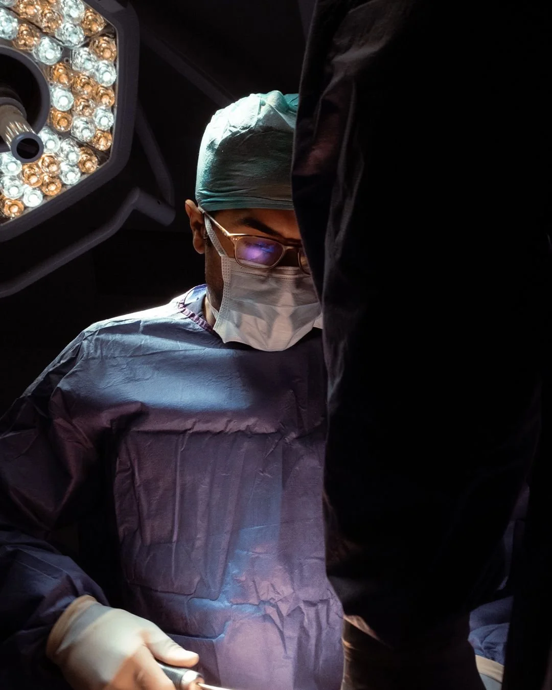 Husain AlQattan. Surgeon wearing glasses, a face mask, a surgical cap, and a gown performing surgery in an operating room with surgical lights overhead.
