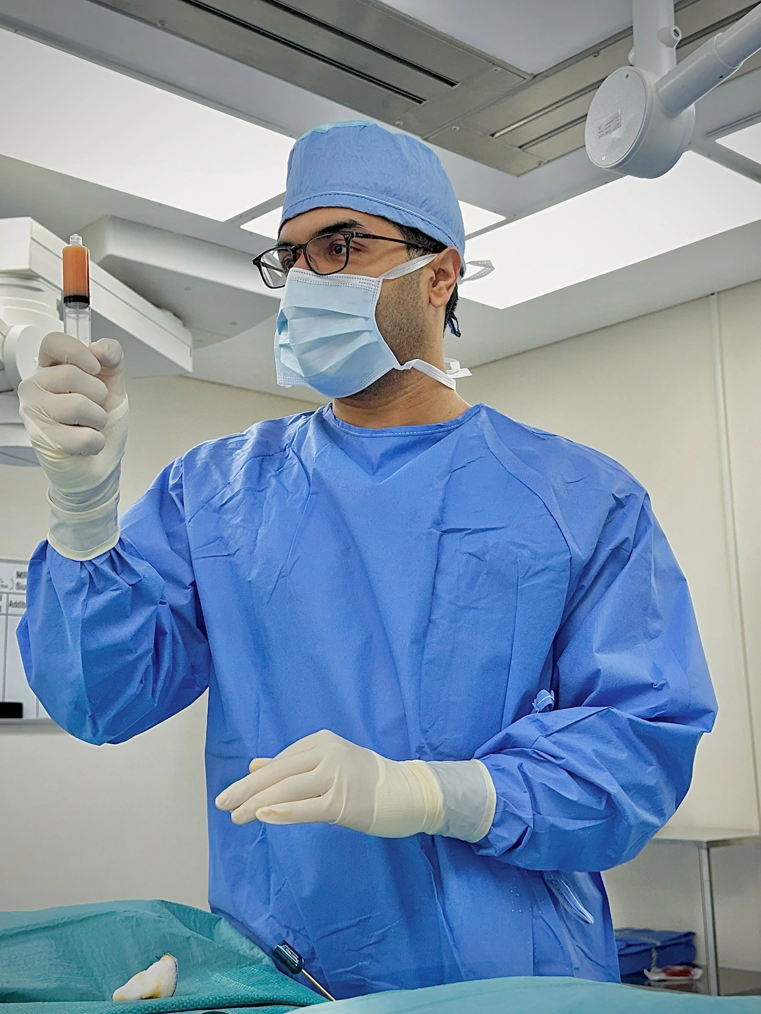 A medical professional wearing a blue surgical gown, gloves, a face mask, glasses, and a blue surgical cap holding a test tube with an orange liquid inside. The person is in an operating room or medical facility.