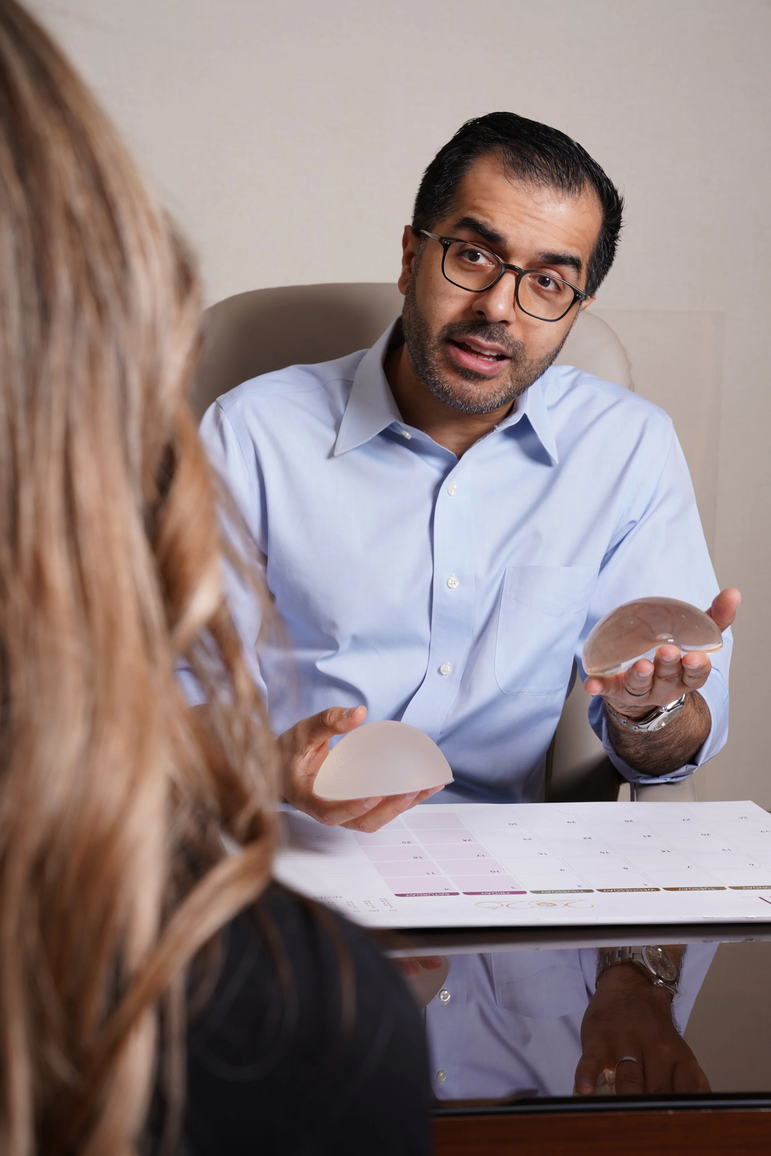 A man in glasses and a light blue shirt holding a breast implant and explaining something to a woman in a professional setting.