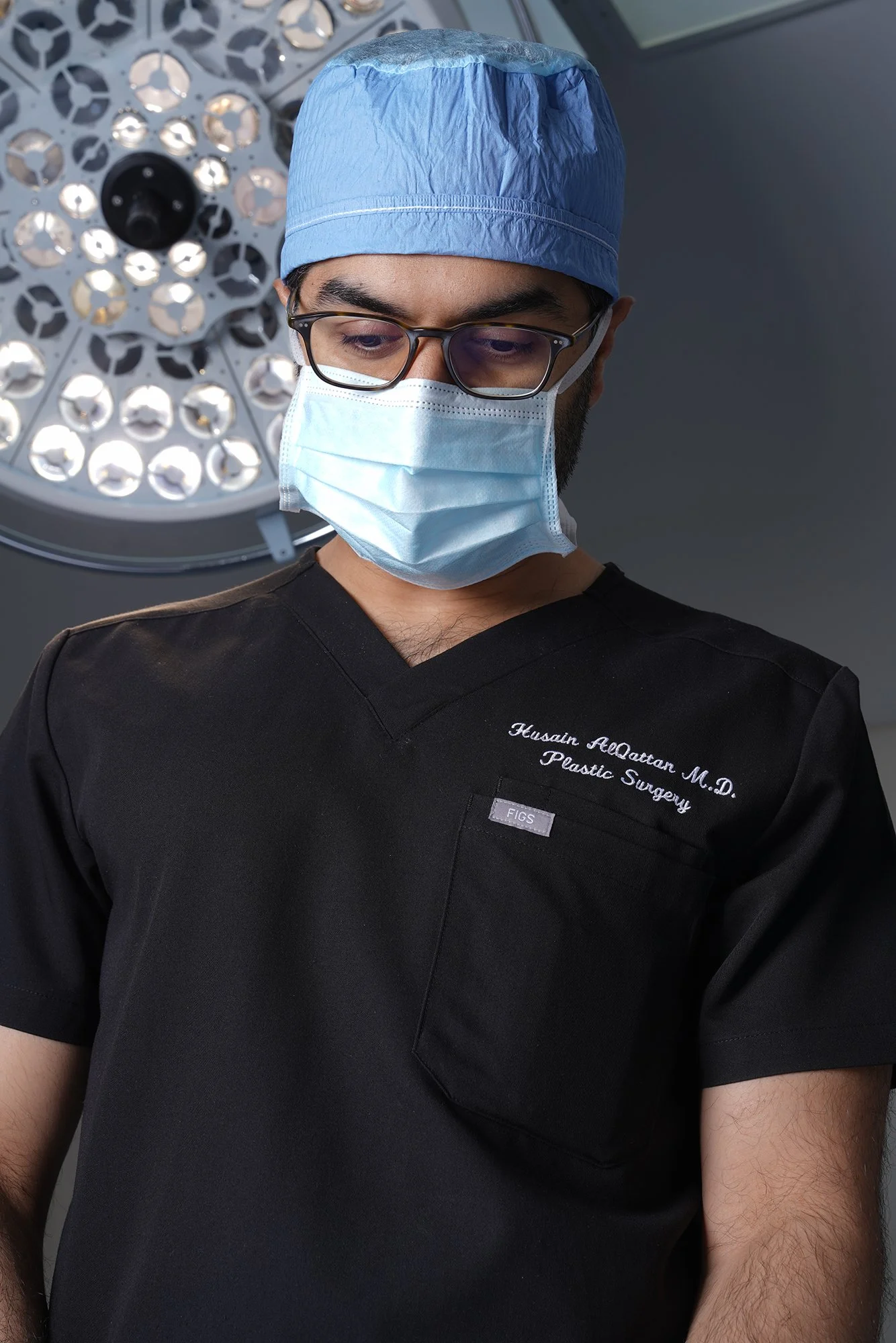 Husain AlQattan. Male surgeon in scrubs and a surgical cap wearing a face mask looking down in an operating room with surgical lights in the background.