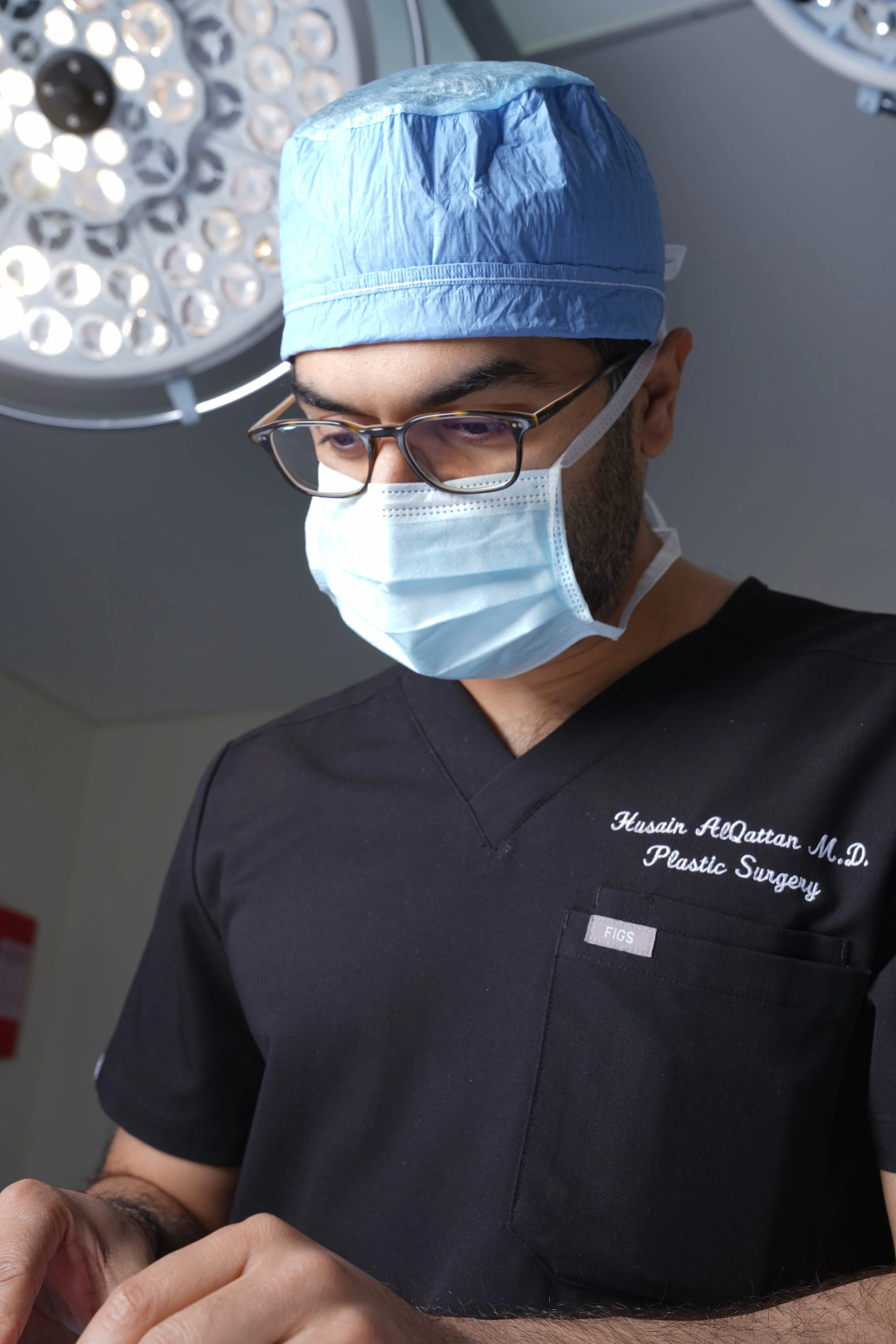 Husain AlQattan. Close-up of a male plastic surgeon wearing a blue surgical cap, glasses, and a face mask inside an operating room with surgical lights overhead.