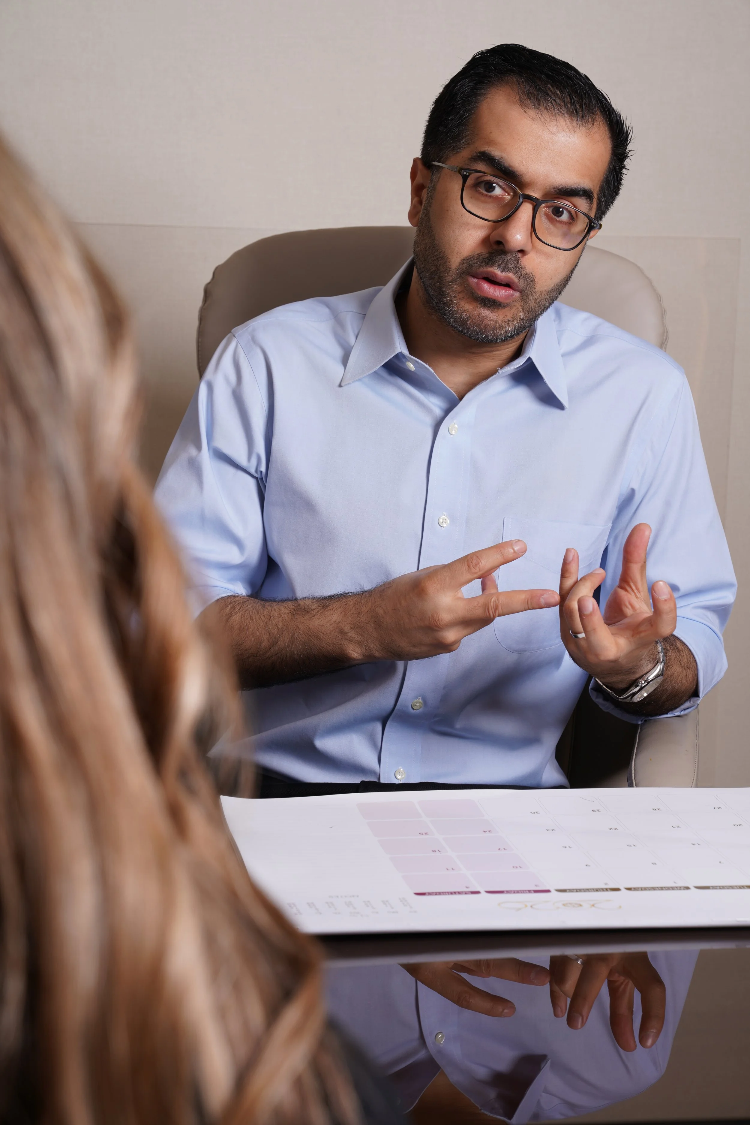 A man with dark hair, glasses, and a beard wearing a light blue dress shirt is sitting at a table, seemingly in a conversation with a woman in front of him, who has long, reddish hair. There is a printed calendar or schedule on the table.