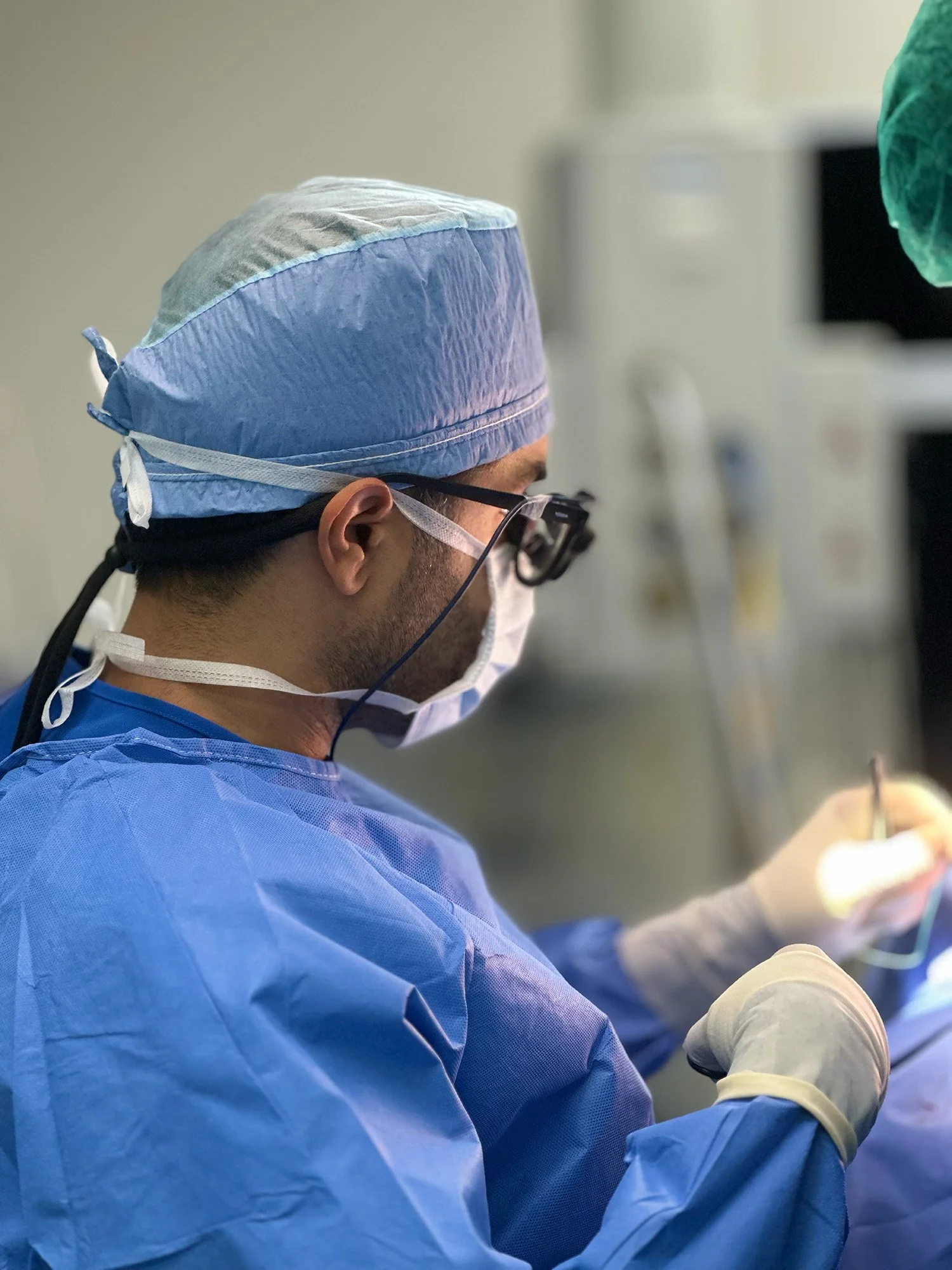 A male surgeon dressed in blue scrubs, wearing a surgical cap, mask, and protective glasses, performing a procedure in a medical setting.