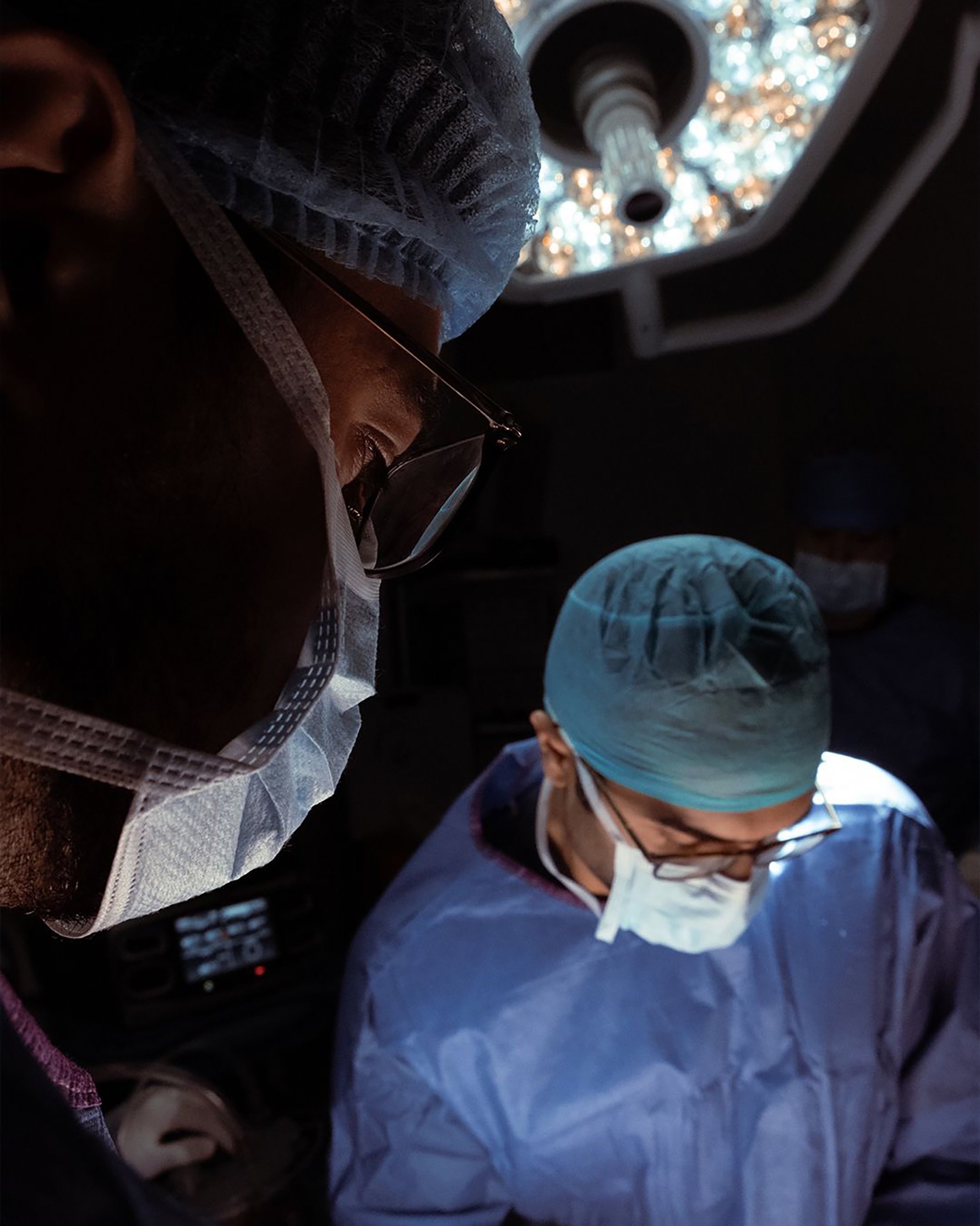 Two medical professionals wearing surgical masks and caps, working together with medical equipment in a dimly lit environment.