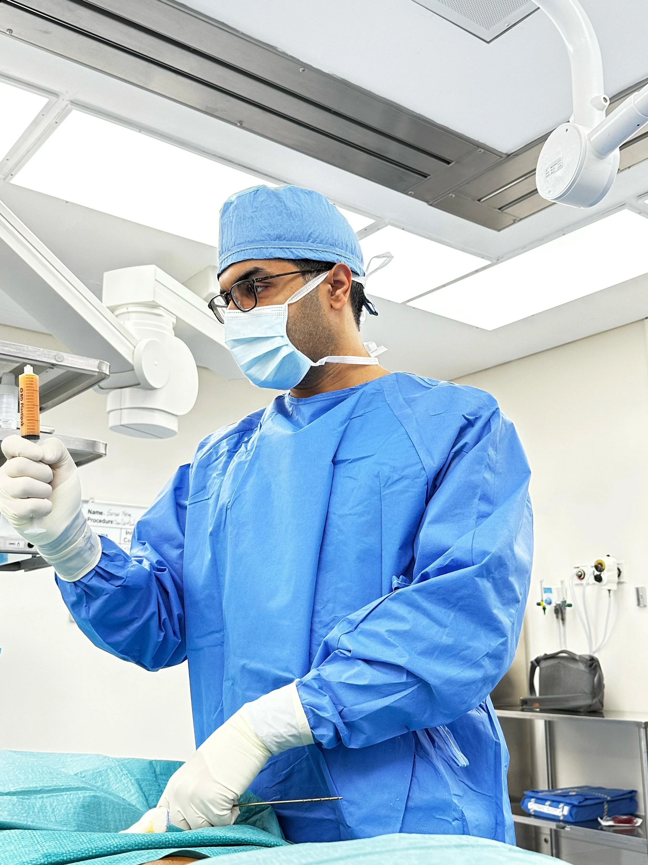 Medical professional wearing a blue surgical gown, mask, gloves, and glasses performing a procedure in an operating room with medical equipment and lights overhead.