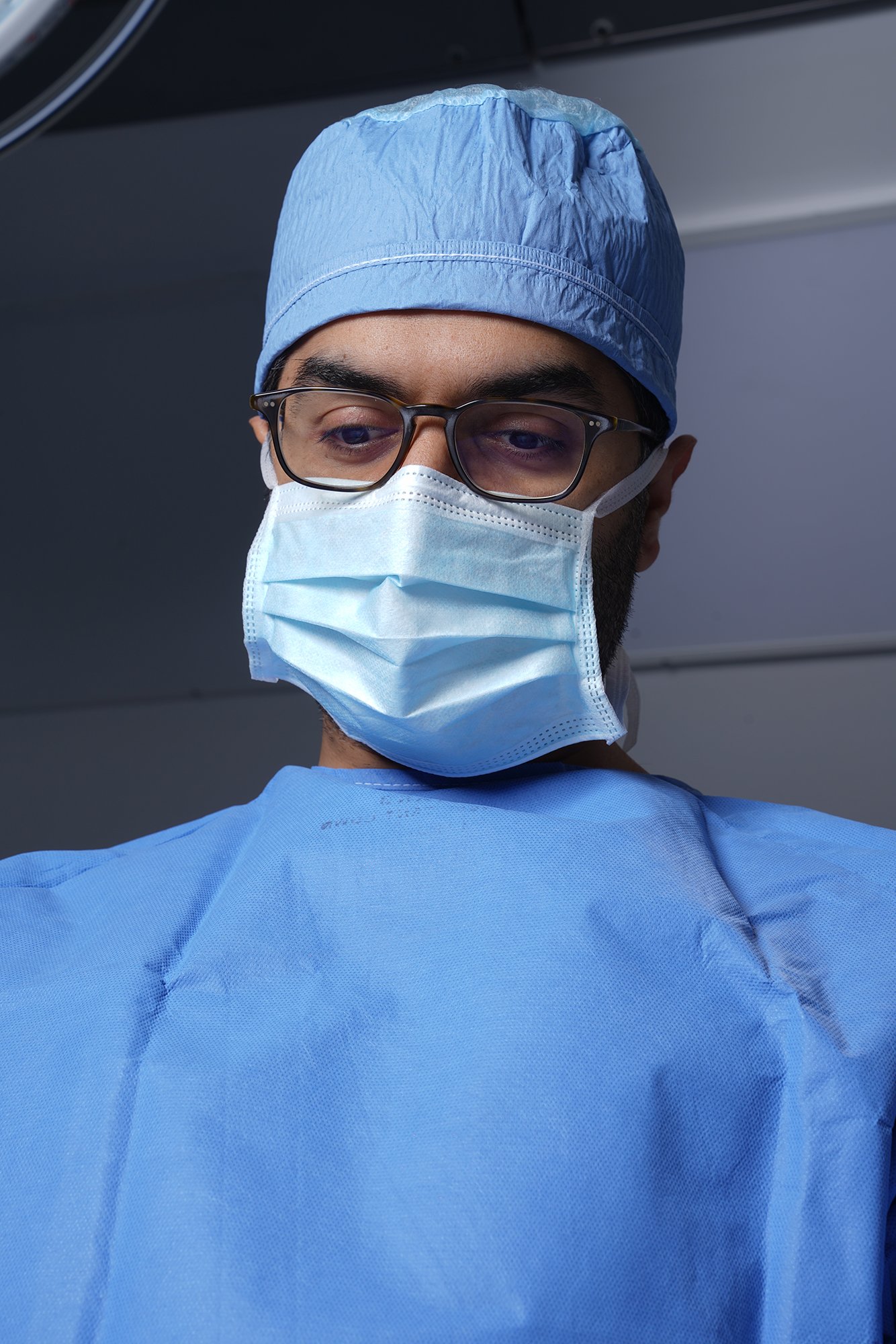 Husain AlQattan. Close-up of a male healthcare worker wearing glasses, a blue surgical mask, and a blue surgical cap, looking downward against a gray background.