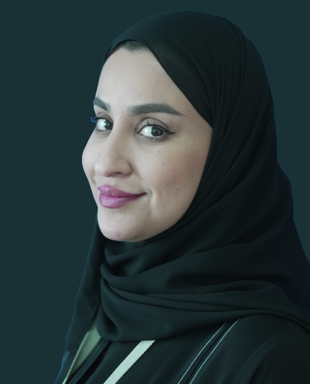 A woman wearing a black hijab and smiling at the camera, with a dark plain background.