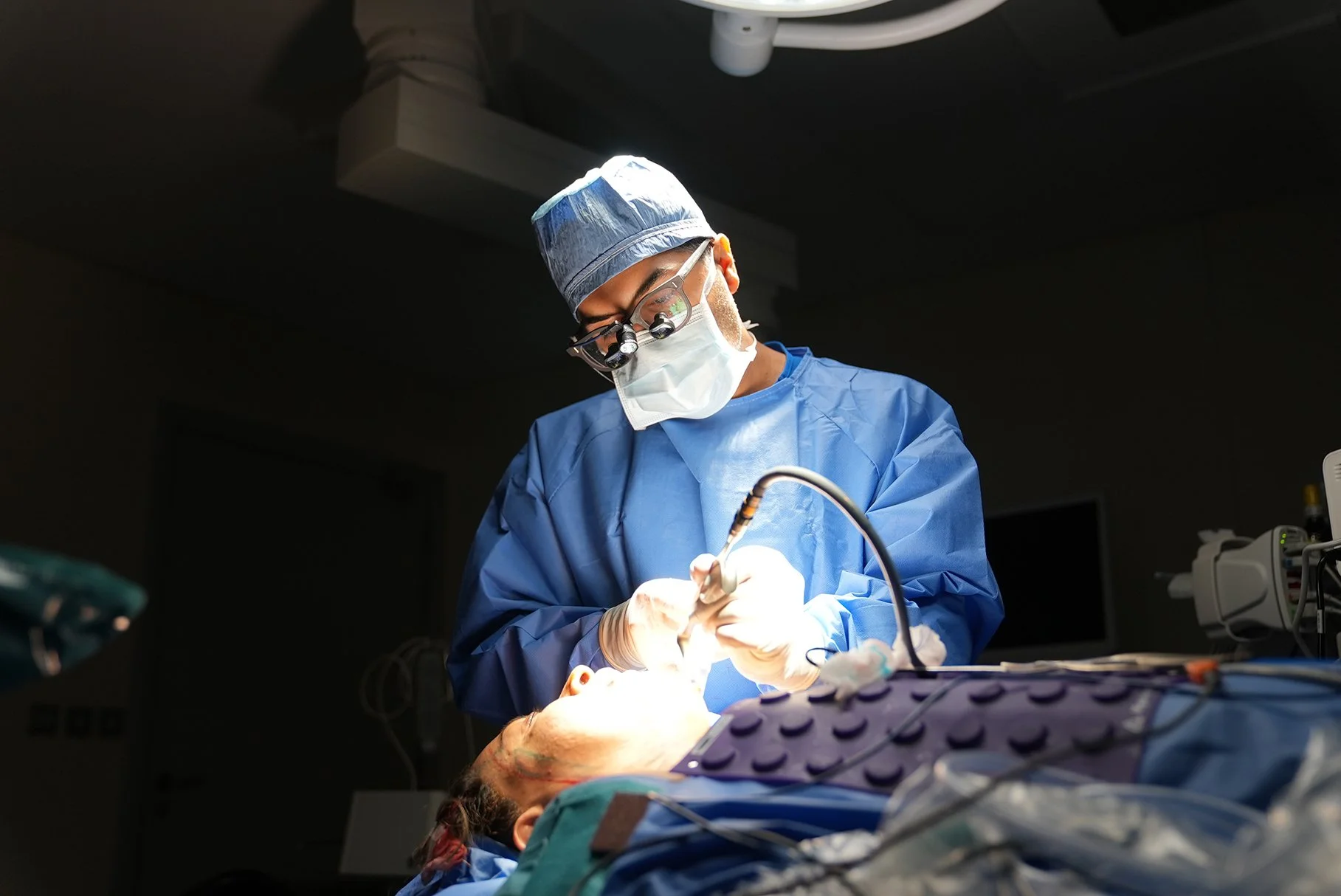 Husain AlQattan. Surgeon performing surgery in an operating room, wearing surgical scrubs, mask, and head covering, with focused lighting and medical equipment around.