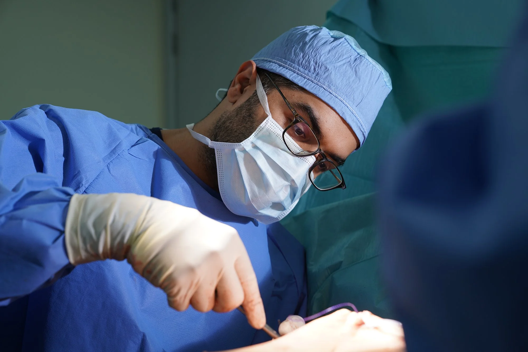 Husain AlQattan. Male surgeon wearing blue surgical scrubs, a cap, glasses, and a face mask looks intently at his work in an operating room.