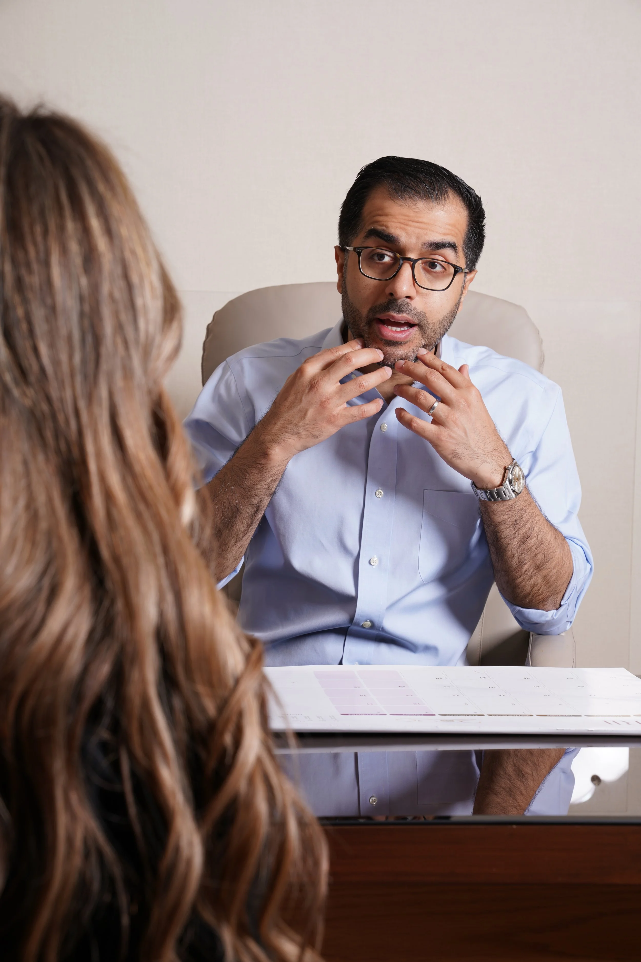A man with glasses and dark hair, wearing a light blue shirt, is talking to a woman with long wavy brown hair, sitting opposite him at a desk. The man appears to be explaining something, with his hands near his chin.