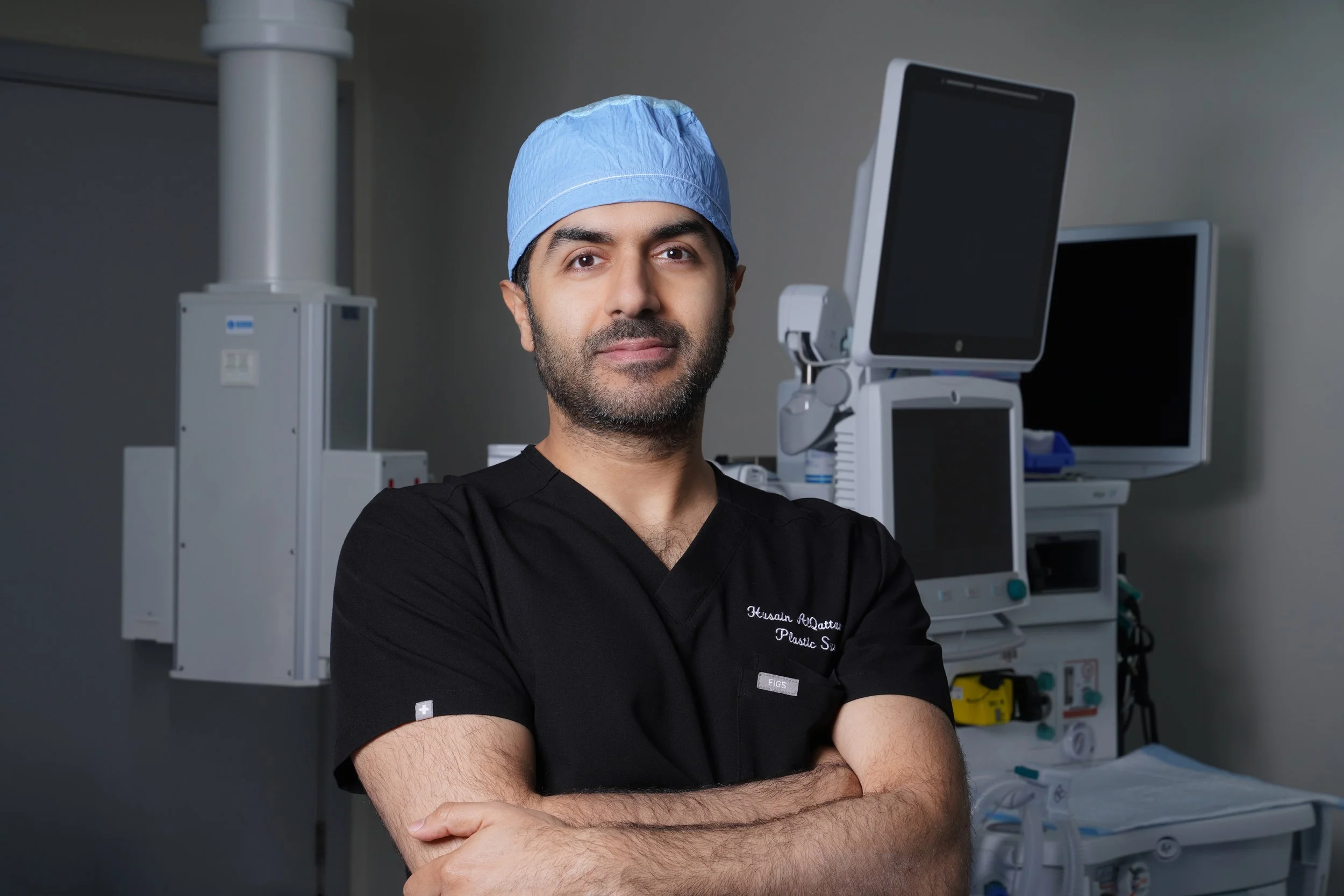 Male surgeon in scrubs and blue surgical cap standing in an operating room with medical monitors and equipment behind him.