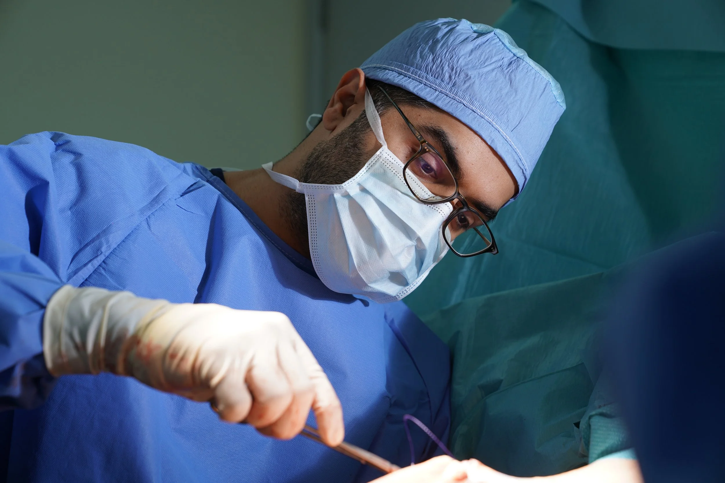 A male surgeon wearing blue scrubs, glasses, and a face mask performing surgery.