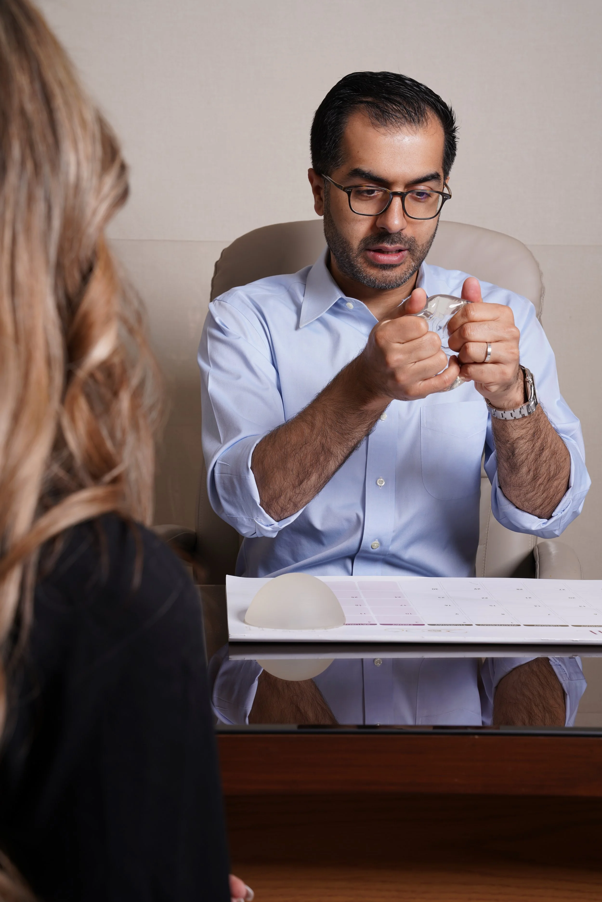 A man with dark hair, glasses, and a beard, wearing a light blue shirt, is sitting at a table and examining a small, clear object. A woman with long, wavy, light brown hair and dark clothing is sitting opposite him, facing away from the camera. There is a sheet of paper with a color chart on the table, and a round white object in front of them.