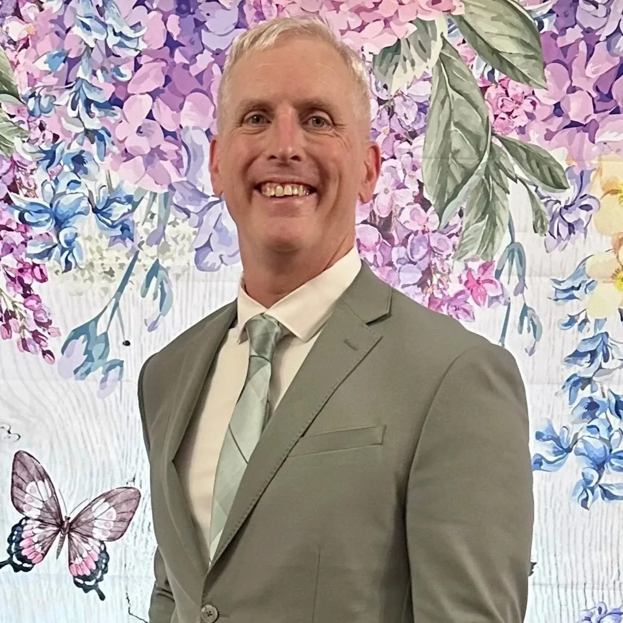 A man in a gray suit and tie smiling in front of a floral and butterfly background.