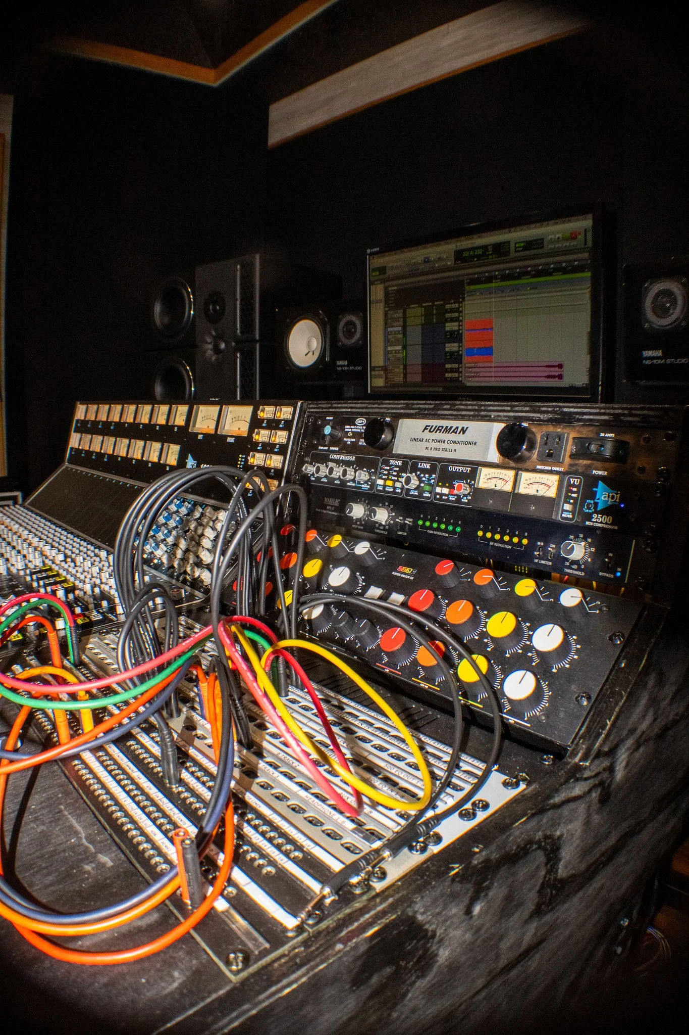 Recording studio mixing console with colorful cables, audio equipment, monitors, and a computer screen displaying audio editing software.