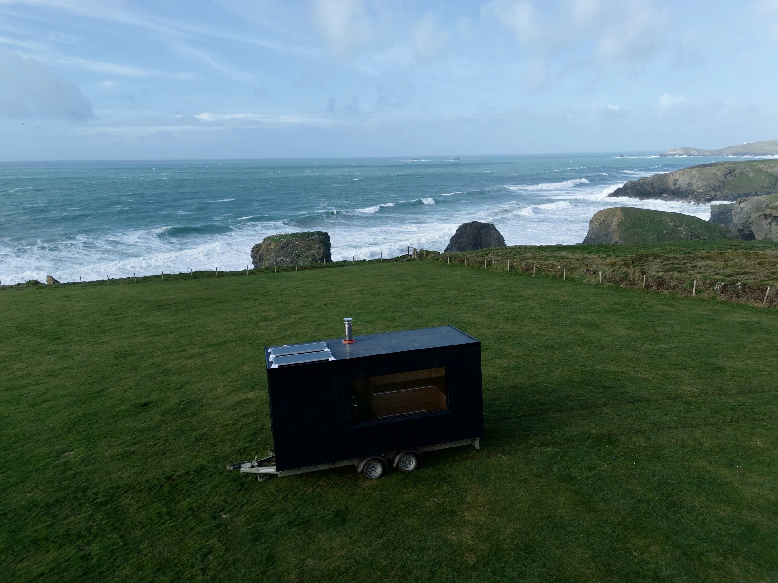 A large green lawn by the ocean with rocks and cliffs in the background, and a black trailer or chassis with solar panels on top in the foreground.