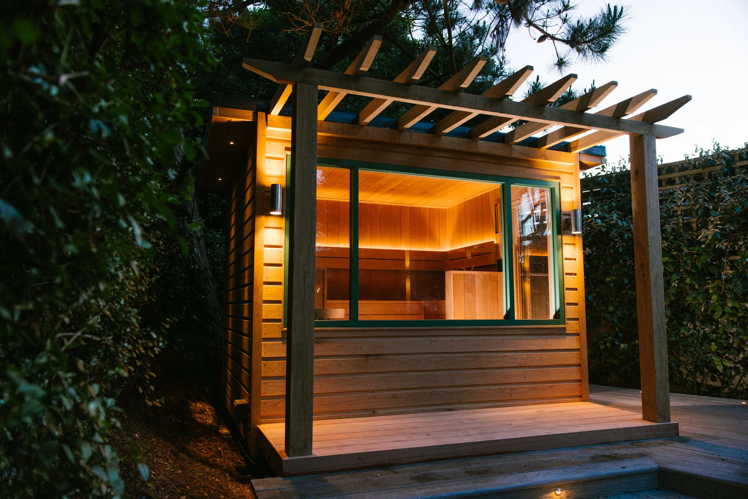 A modern wooden sauna with glass doors illuminated from within, surrounded by lush greenery and trees as evening falls.