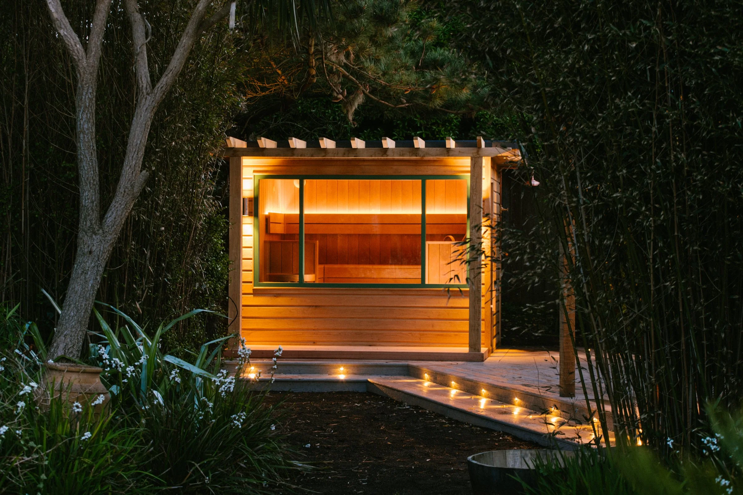 A small wooden structure with a sliding glass door and warm lighting, surrounded by lush greenery and trees in a garden setting at night.