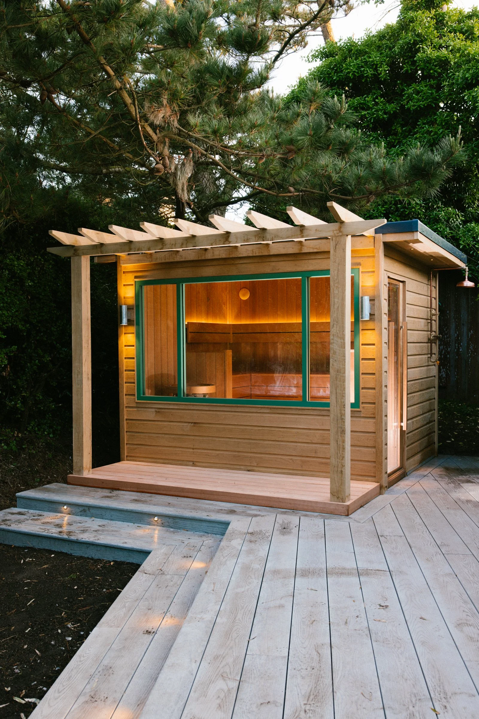 Small wooden outdoor structure with a slanted roof, large windows, surrounded by trees and a light-colored wooden deck with built-in lighting.