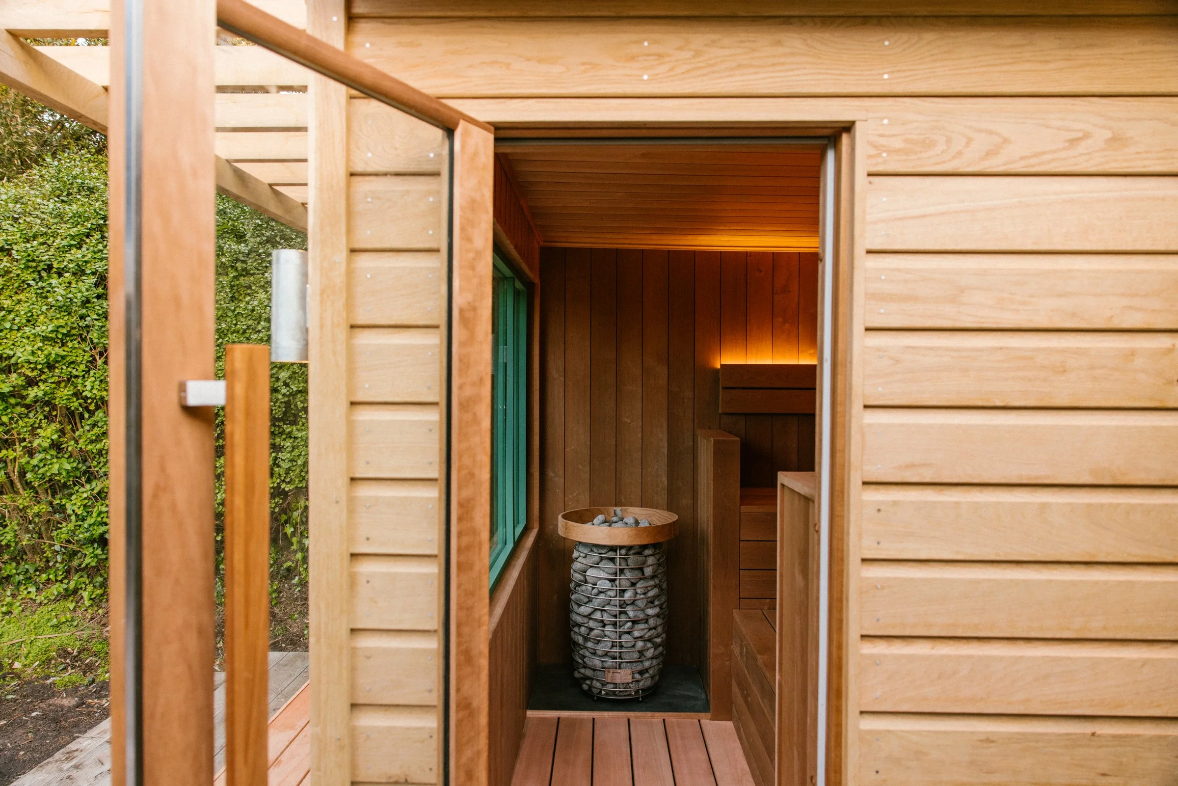 Inside a small, modern, wooden sauna with wooden walls, ceiling, and floor, featuring a basket of smooth stones in the center and a warm light illuminating the interior from the back corner.