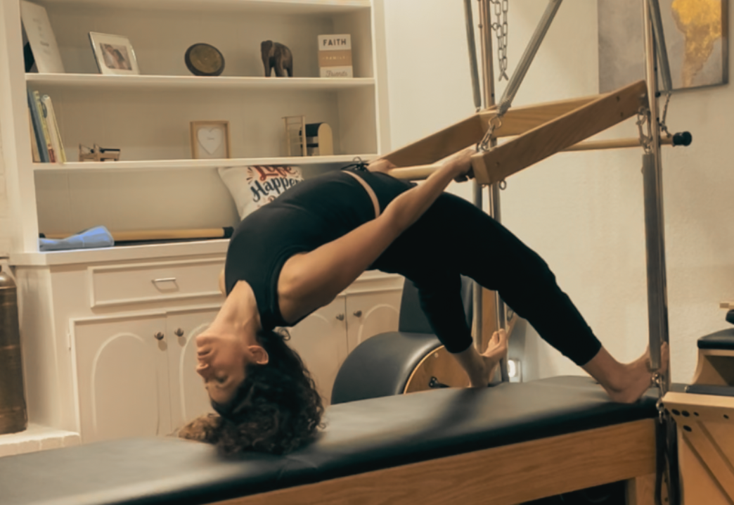 A woman performing a yoga pose on a Pilates reformer in a home gym, with shelves and framed photos in the background.