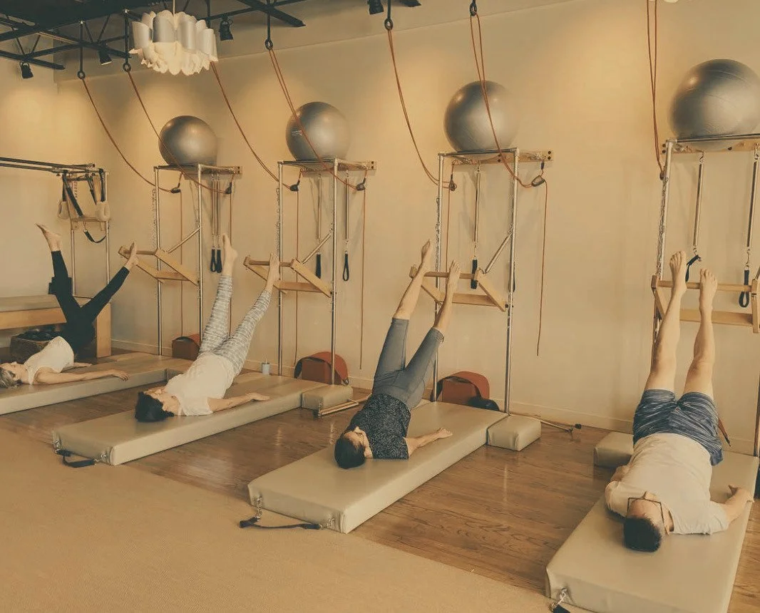 Four individuals are lying on their backs on mats in a Pilates studio, with their legs elevated and supported by wooden bars attached to the wall. Large exercise balls are suspended from the ceiling above each person.