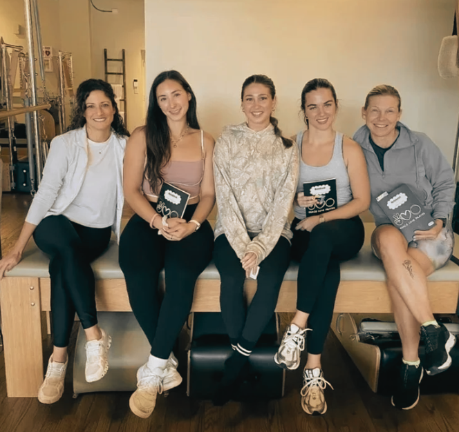 Five women sitting on a padded bench in a fitness or physical therapy room, smiling at the camera. They are holding signs, and there are exercise equipment and a ladder in the background.