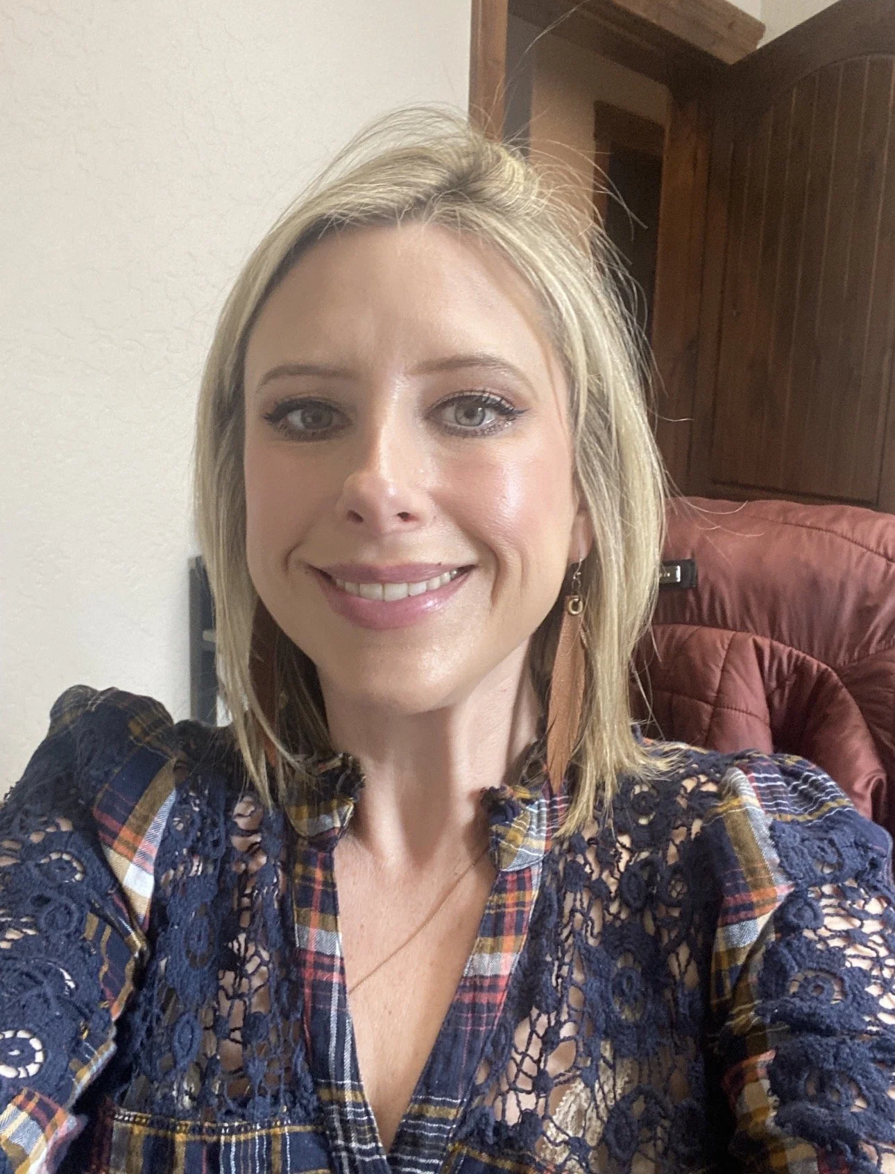 A young woman with blonde hair and blue eyes smiling at the camera, wearing a plaid shirt with lace details, sitting in a room with wooden furniture and a red jacket in the background.