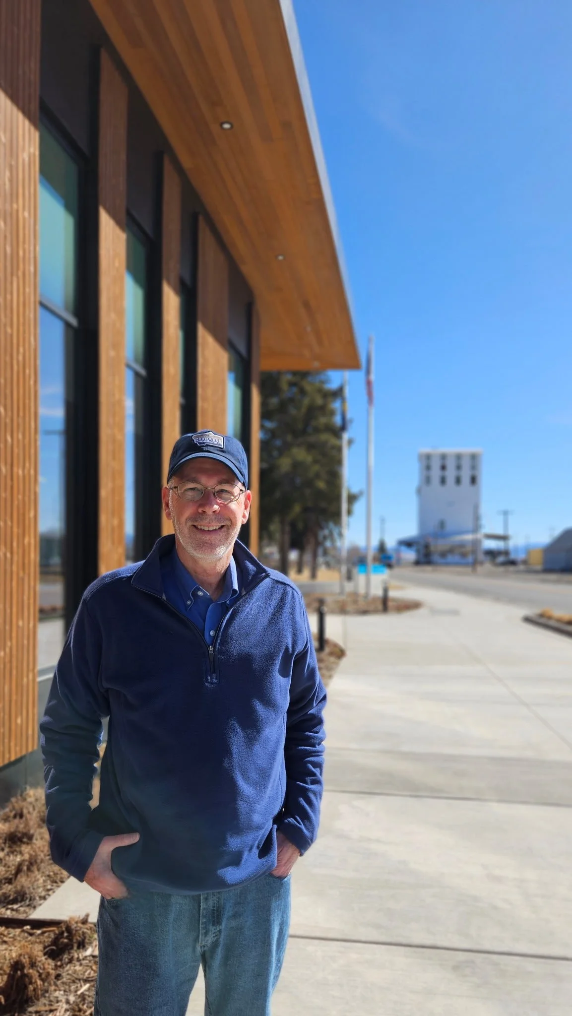 Smiling man wearing a blue jacket, blue cap, and glasses standing outdoors near a modern wooden building with large windows on a sunny day.