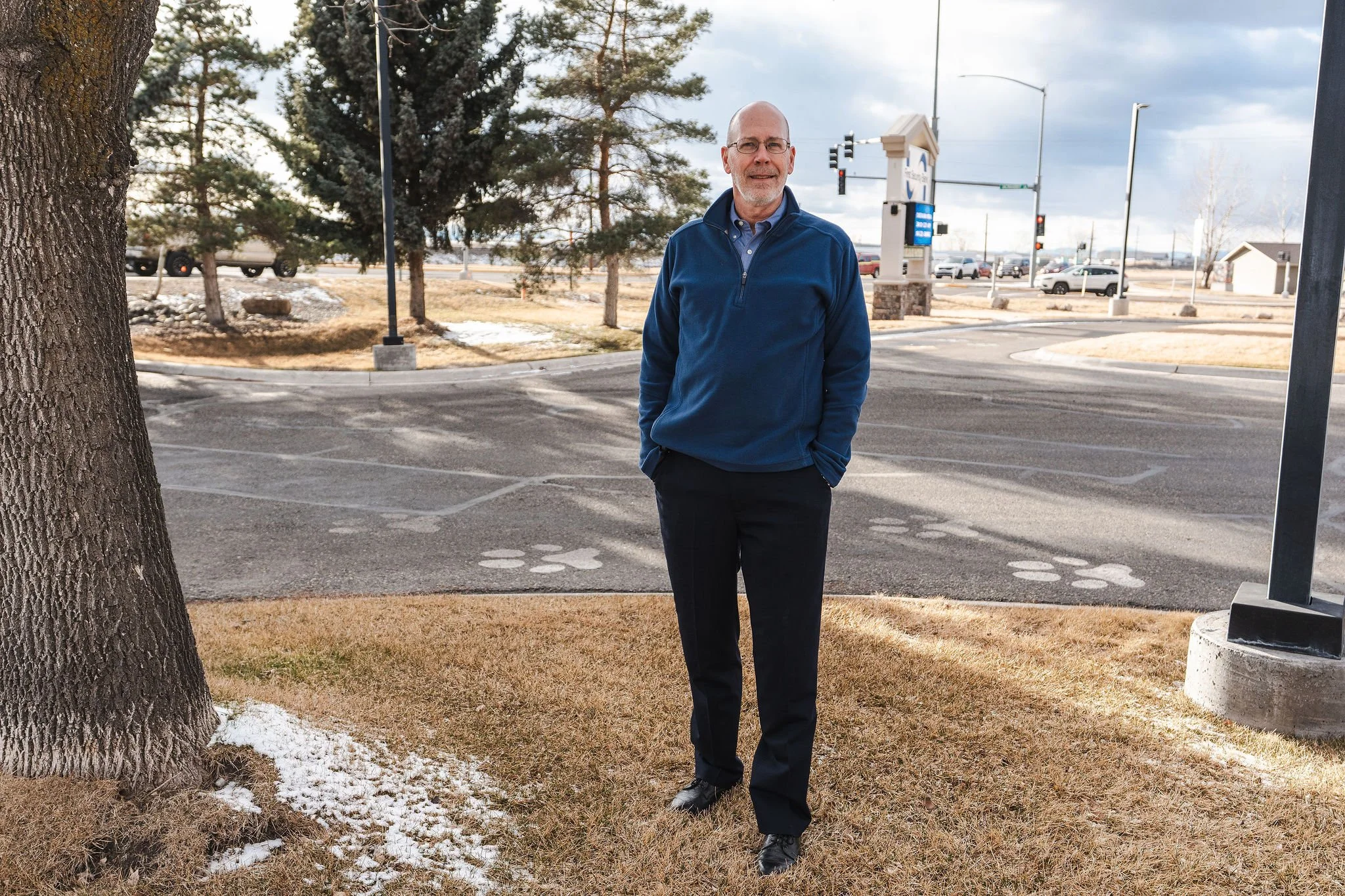 A man with glasses, a beard, and a bald head walking outdoors near a parking lot on a cloudy day. He is wearing a blue quarter-zip jacket, dark pants, and black shoes. There are trees, some snow patches, and cars in the background.