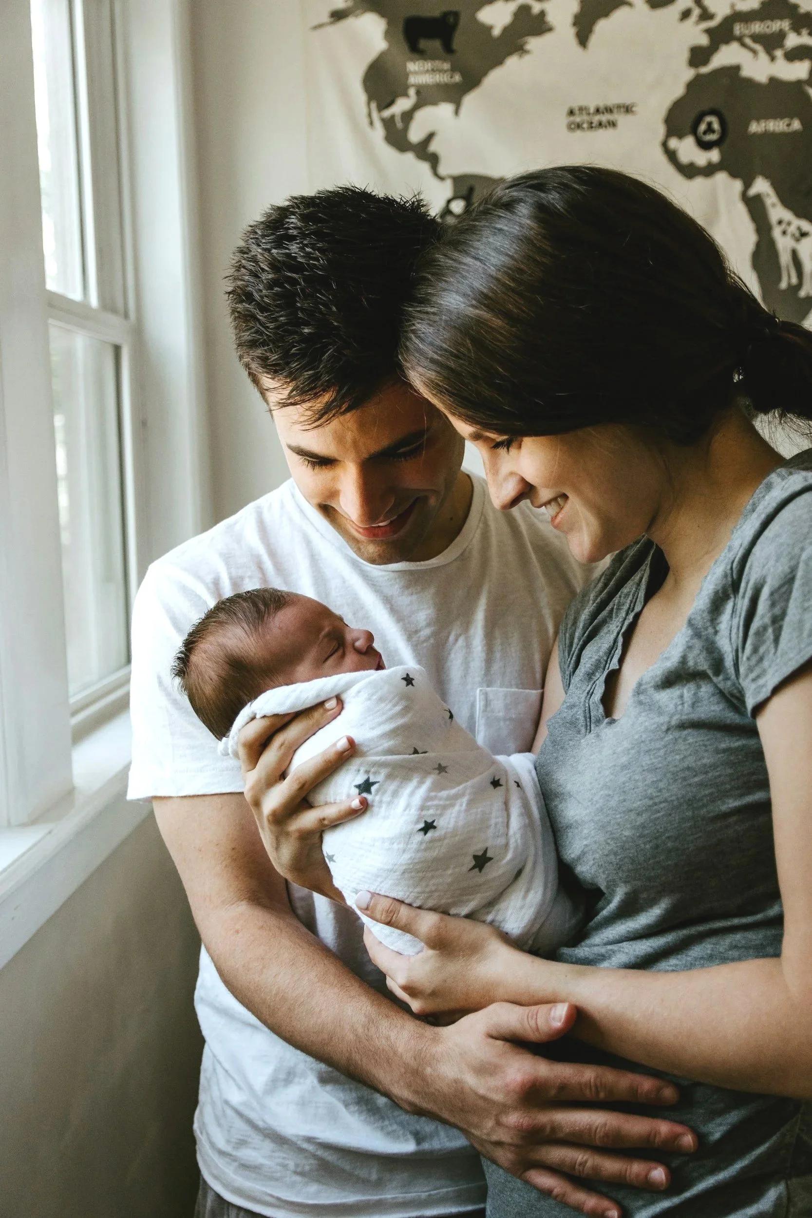 A young couple holding a newborn baby near a window, smiling and looking at the baby affectionately.