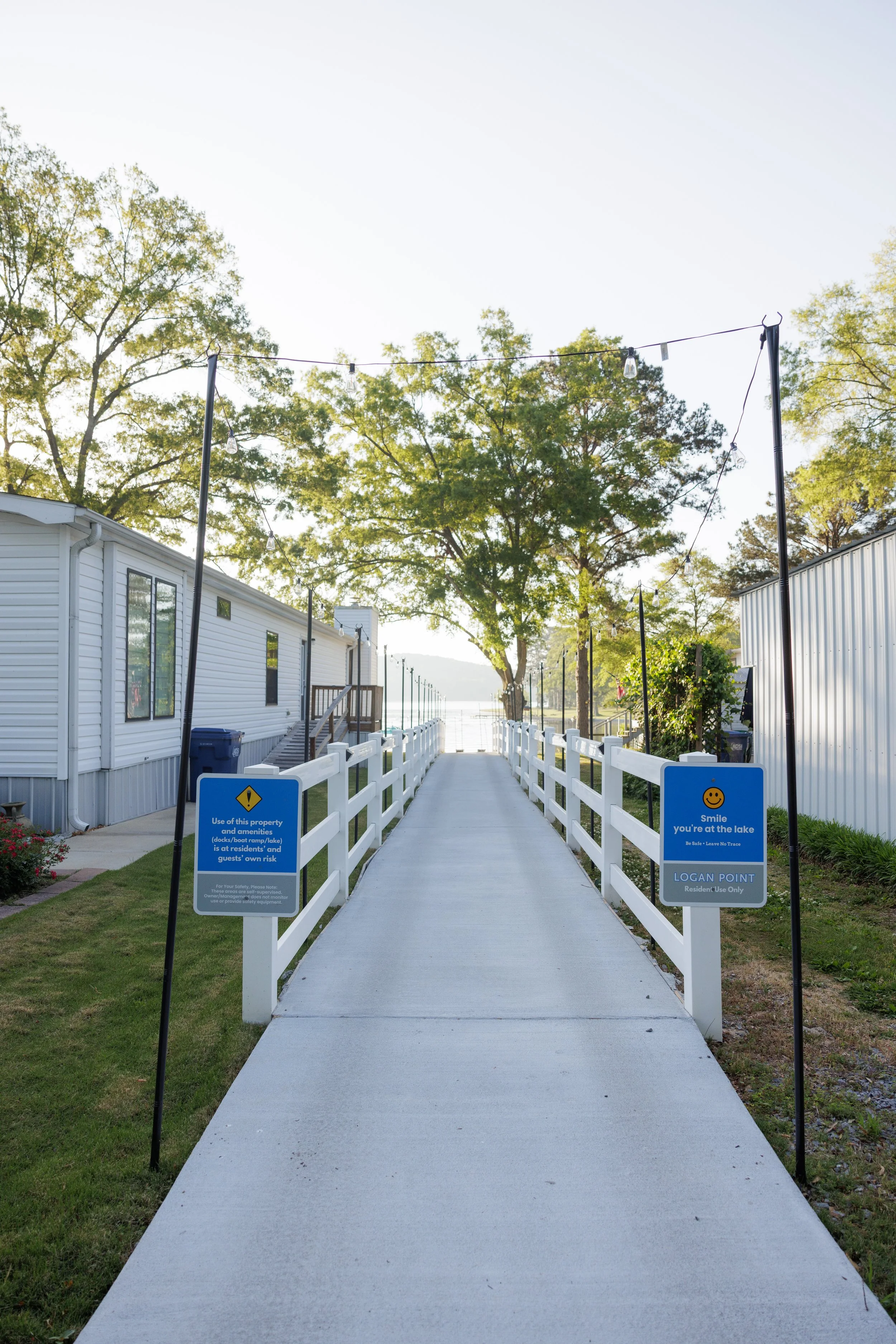 A concrete pathway with white fencing on each side leads towards a lake, lined with trees and buildings; signs warning residents to use the area at their own risk and emphasizing smiley face positivity at the end of the path.