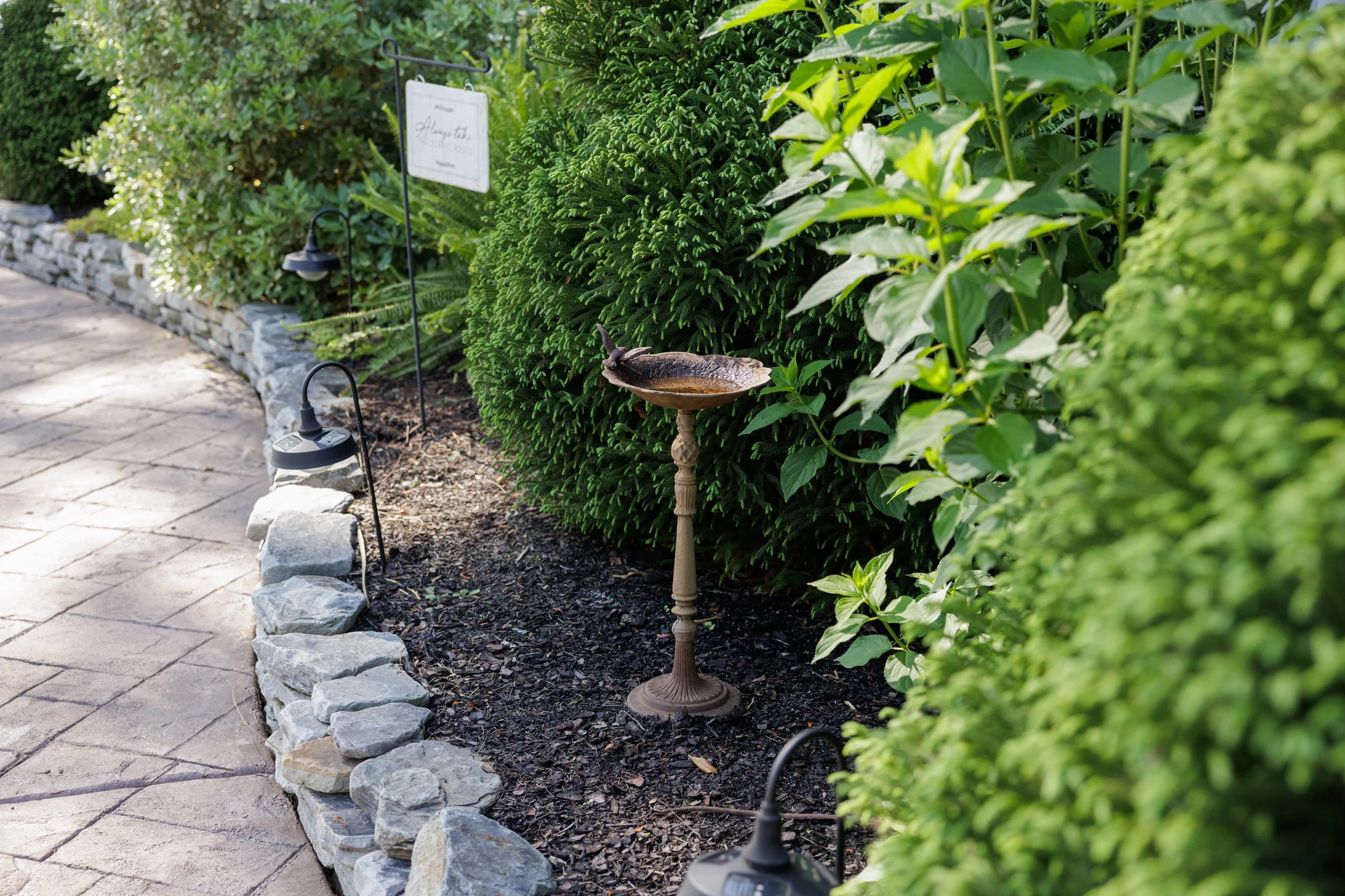 Garden walkway with stone border, surrounded by lush green bushes and plants, with a birdbath and outdoor lights.