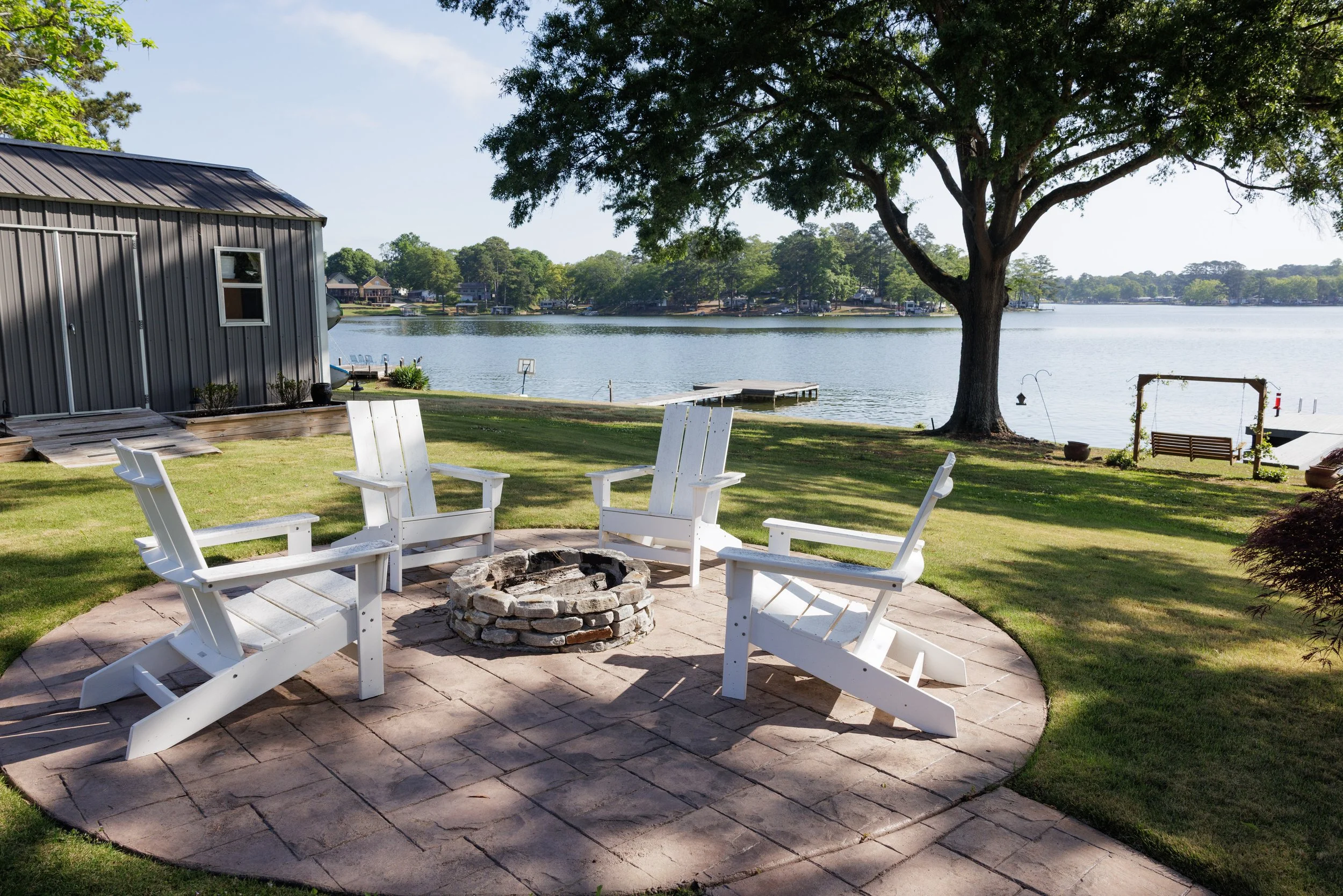 Backyard with a fire pit surrounded by six white chairs on a stone patio. A large tree provides shade, overlooking a lake with houses and boats in the background.