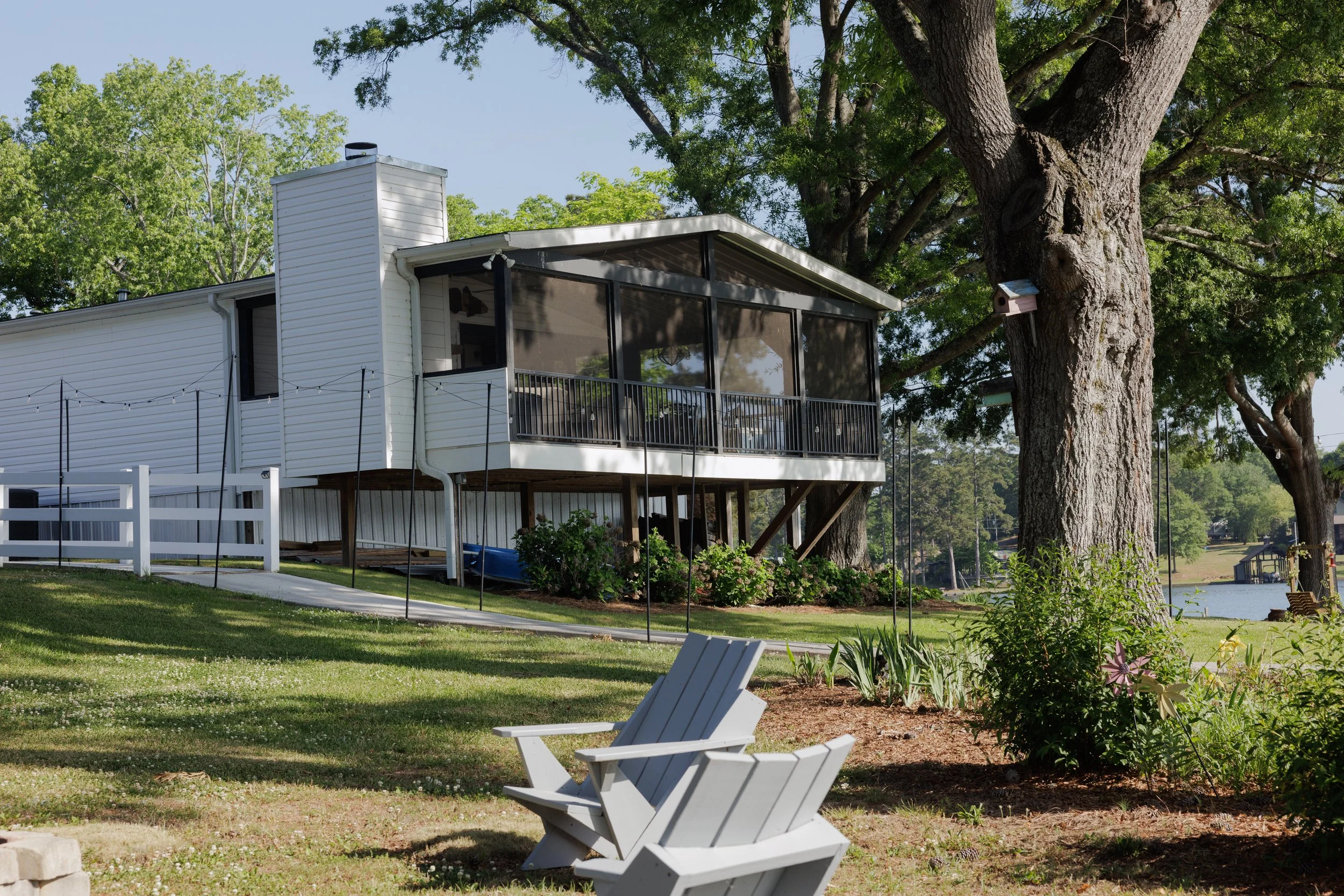 A lakeside yard with a white house on stilts, a deck with black railing, a large tree with a birdhouse, and two white Adirondack chairs in the yard.