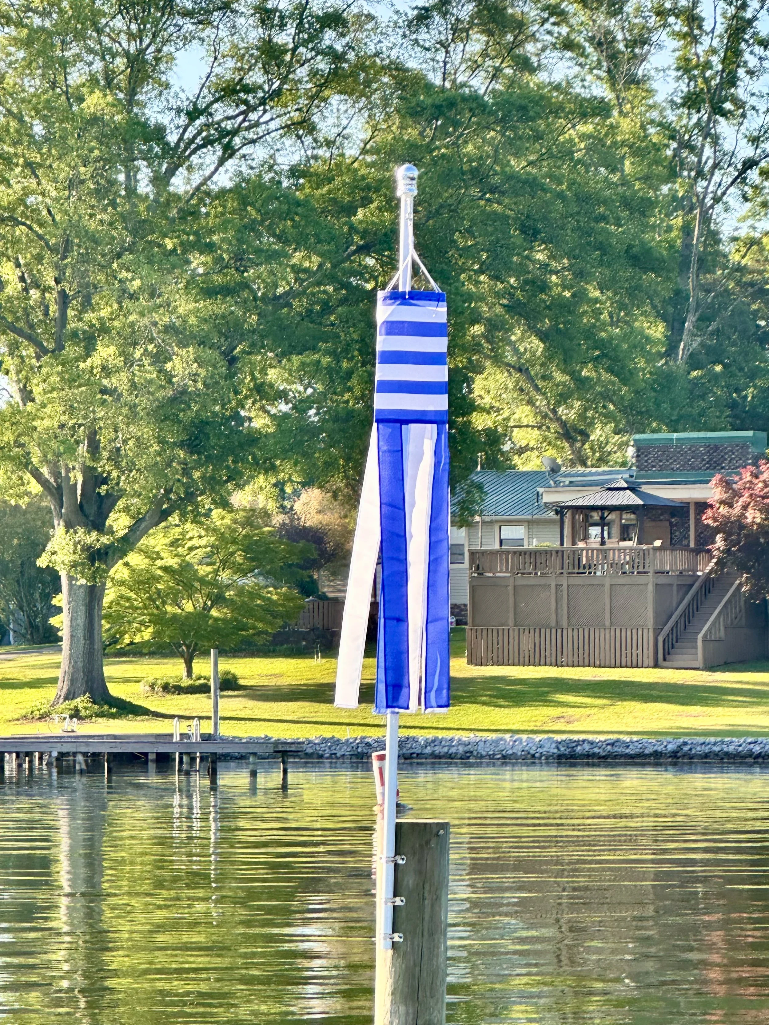 A blue and white striped windsock on a pole near a body of water, with trees and houses in the background.