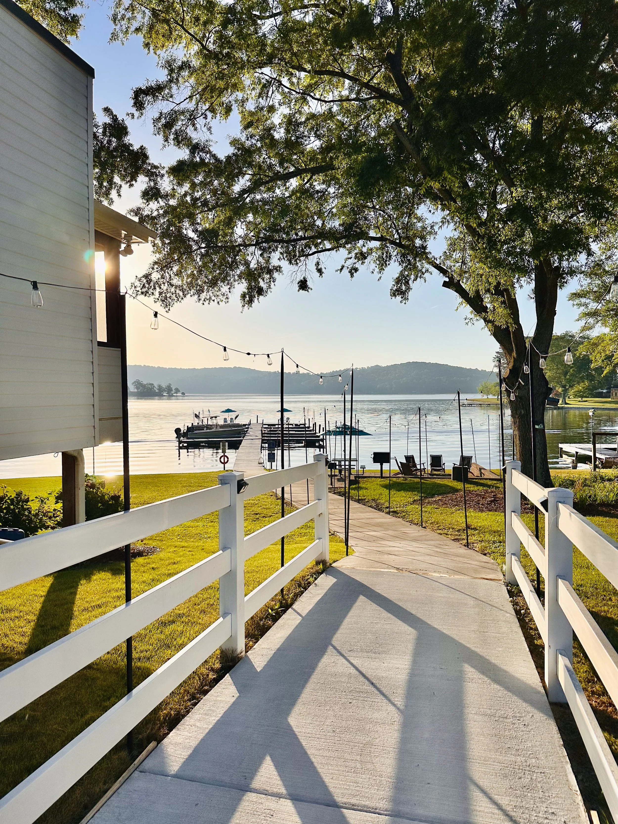 A concrete pathway leads to a dock on a lake with boats, surrounded by green grass and trees, with string lights hanging along the pathway. The sky is clear, and the sun is shining through the tree leaves.