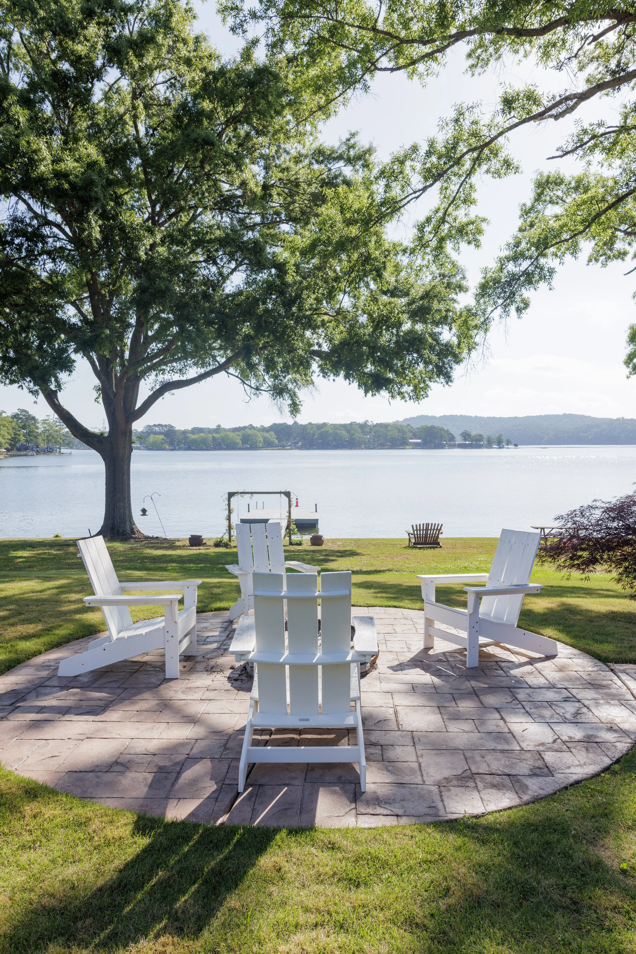 A lakeside backyard patio with white Adirondack chairs arranged in a circle around a fire pit, overlooking the water and a large tree providing shade.