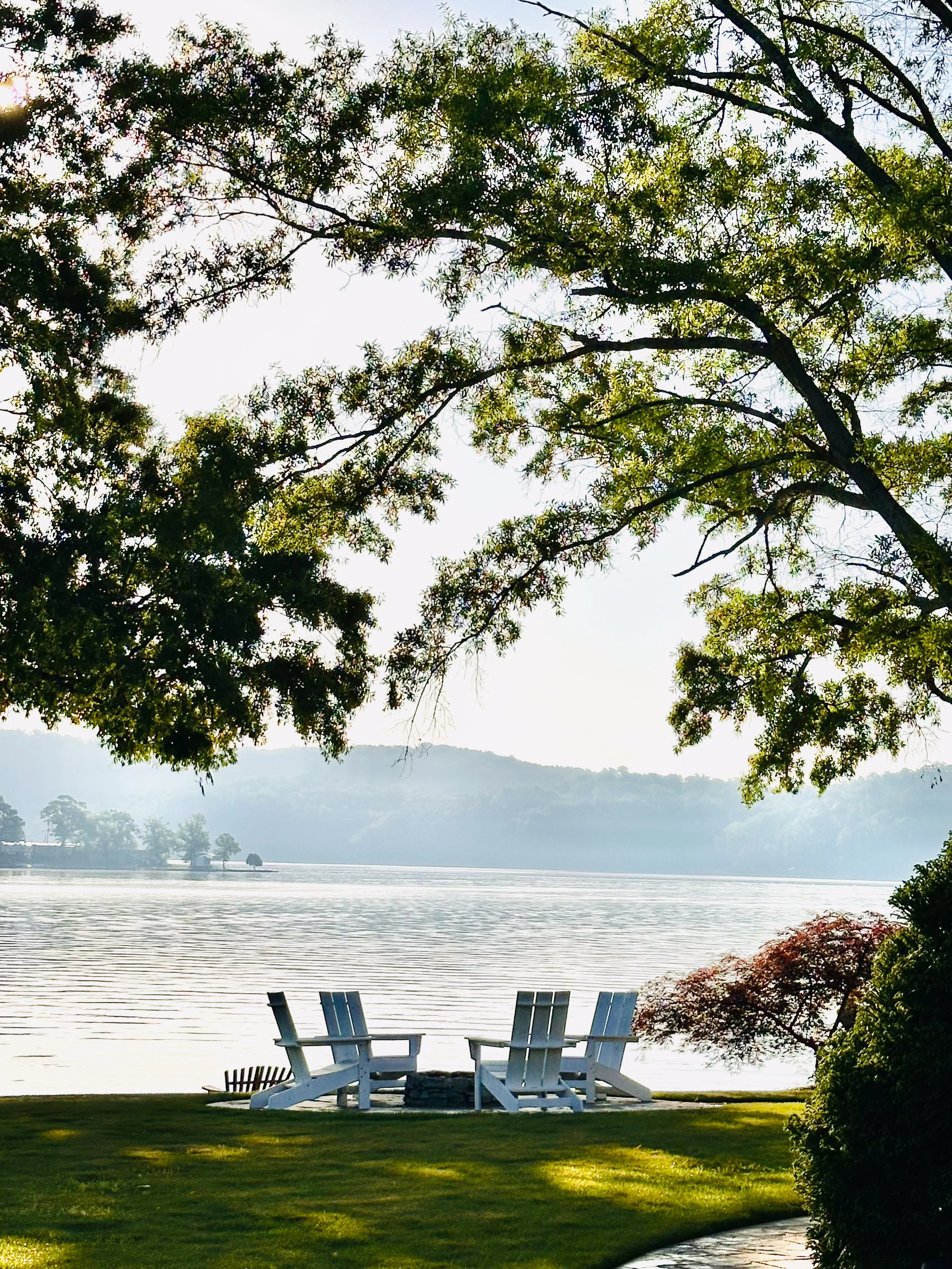Two white Adirondack chairs on a grassy lawn by a lake, with trees arching overhead and mountains in the background.