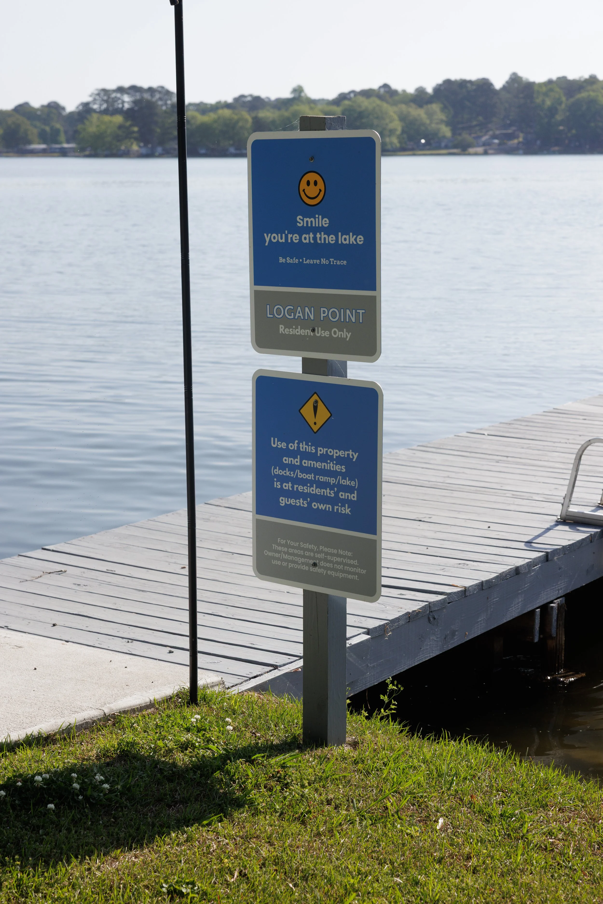 Sign at a lake with a blue background and white text that reads "Smile you're at the lake" with a smiley face icon above. The sign also says "Be Safe - Leave No Trace," and below it, there's another sign with a yellow warning icon and text about prop