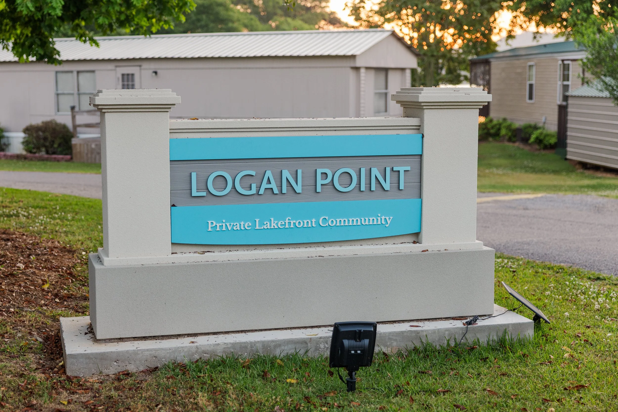 A sign that reads "Logan Point Private Lakefront Community" located outdoors with trees and residential buildings in the background at sunrise or sunset.