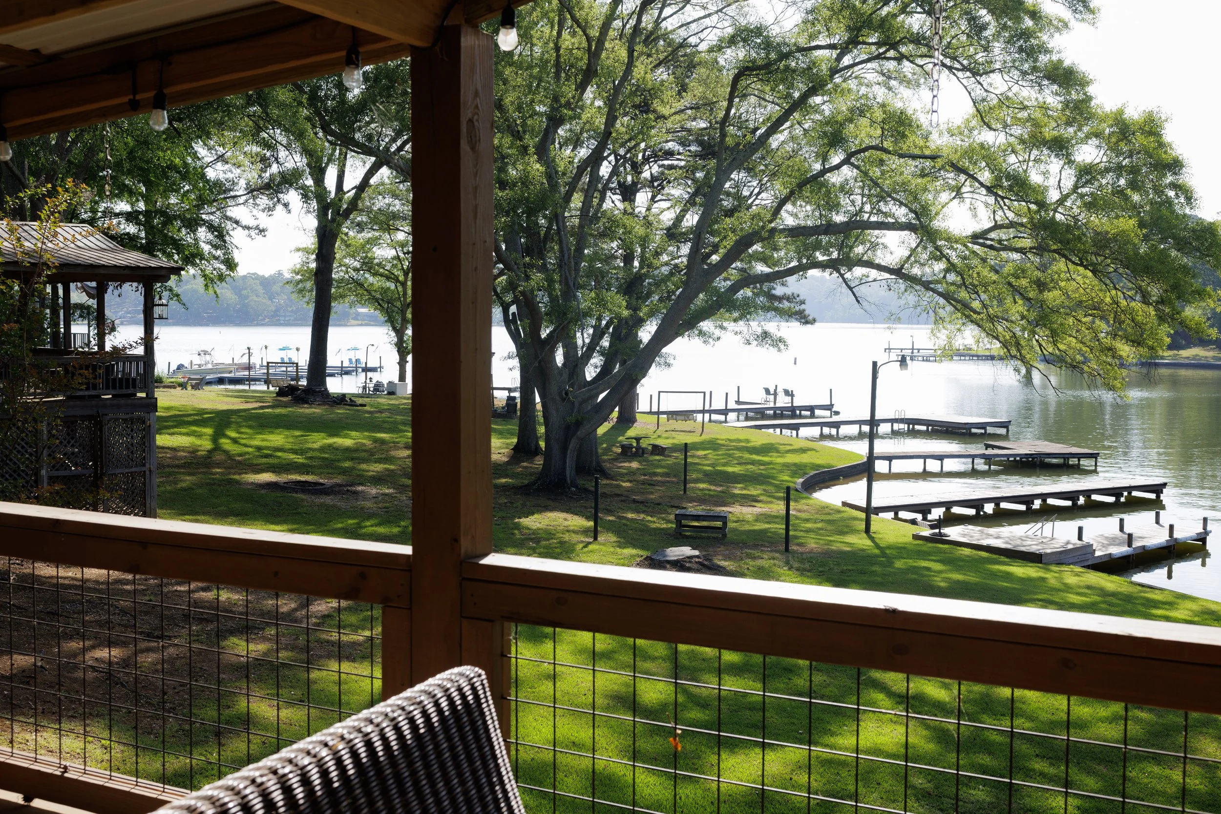 View of a lakeside from a porch with a wooden railing, trees, docks, and water in the background.