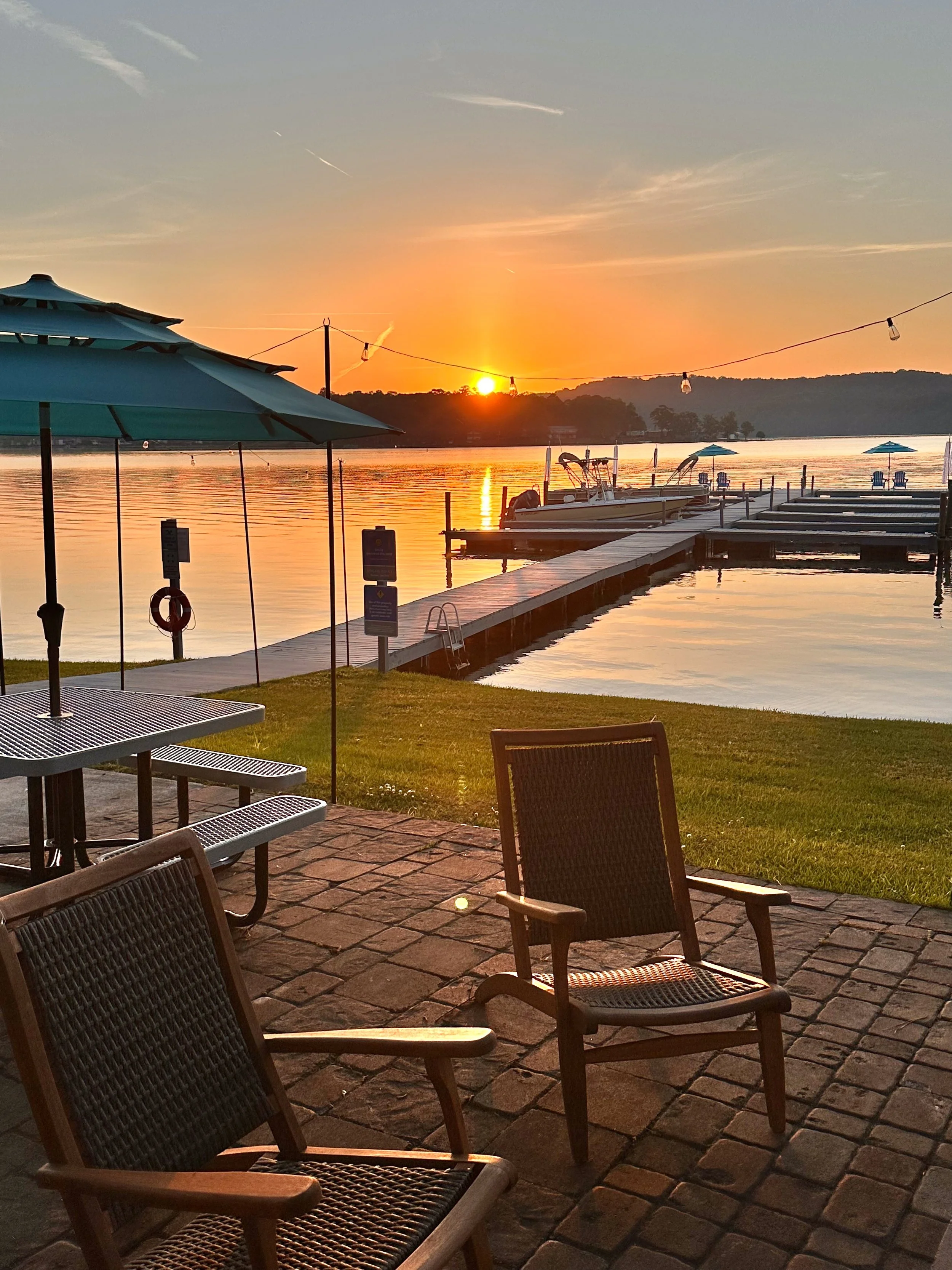 A lakeside dock at sunset with chairs and umbrellas on a patio, boats tied to the dock, calm water, and a distant tree-lined shoreline.