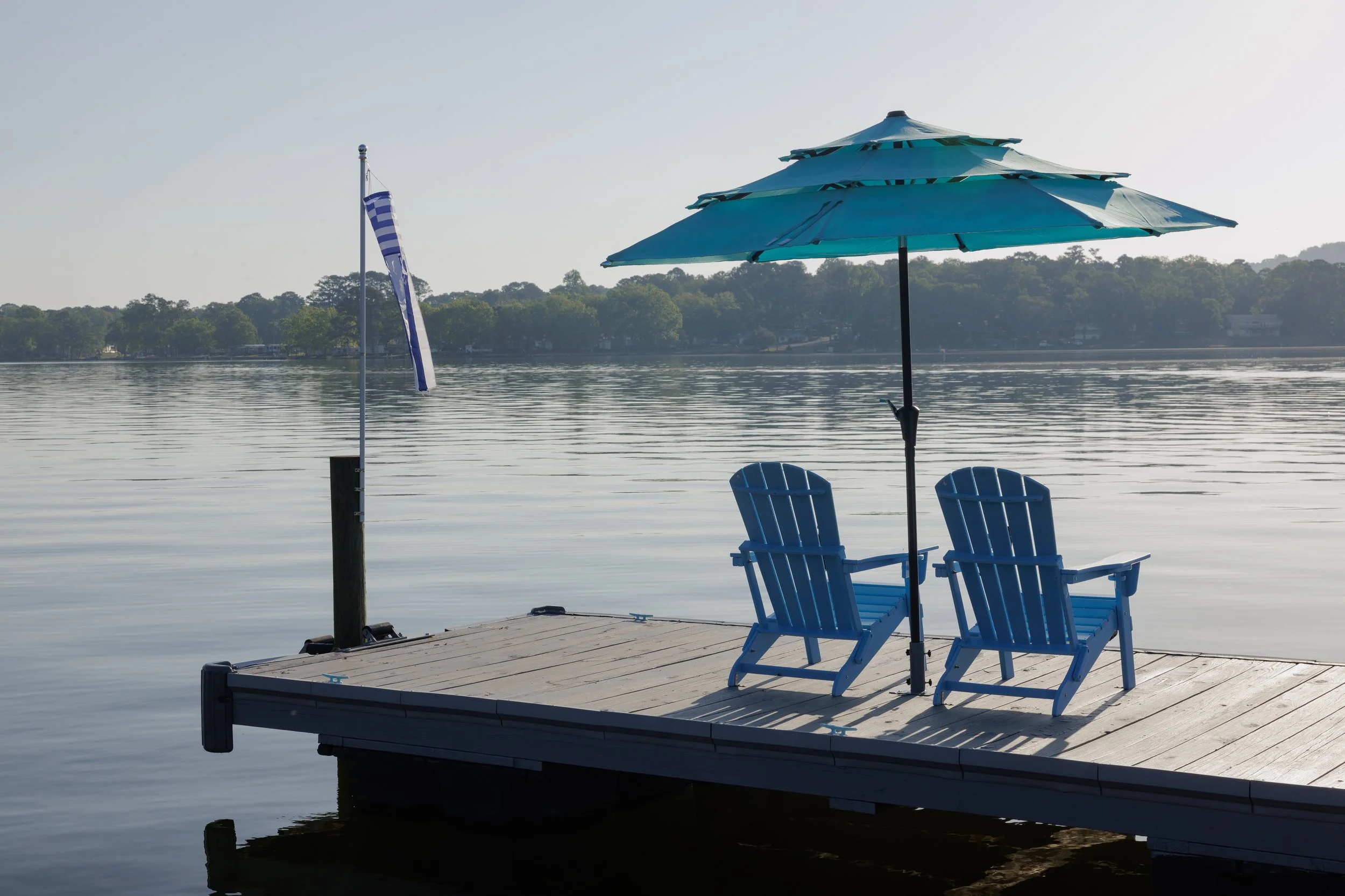 A lakeside dock with two blue Adirondack chairs, a blue patio umbrella, a flagpole with a Greek flag, calm water, and a tree-lined shoreline in the background.