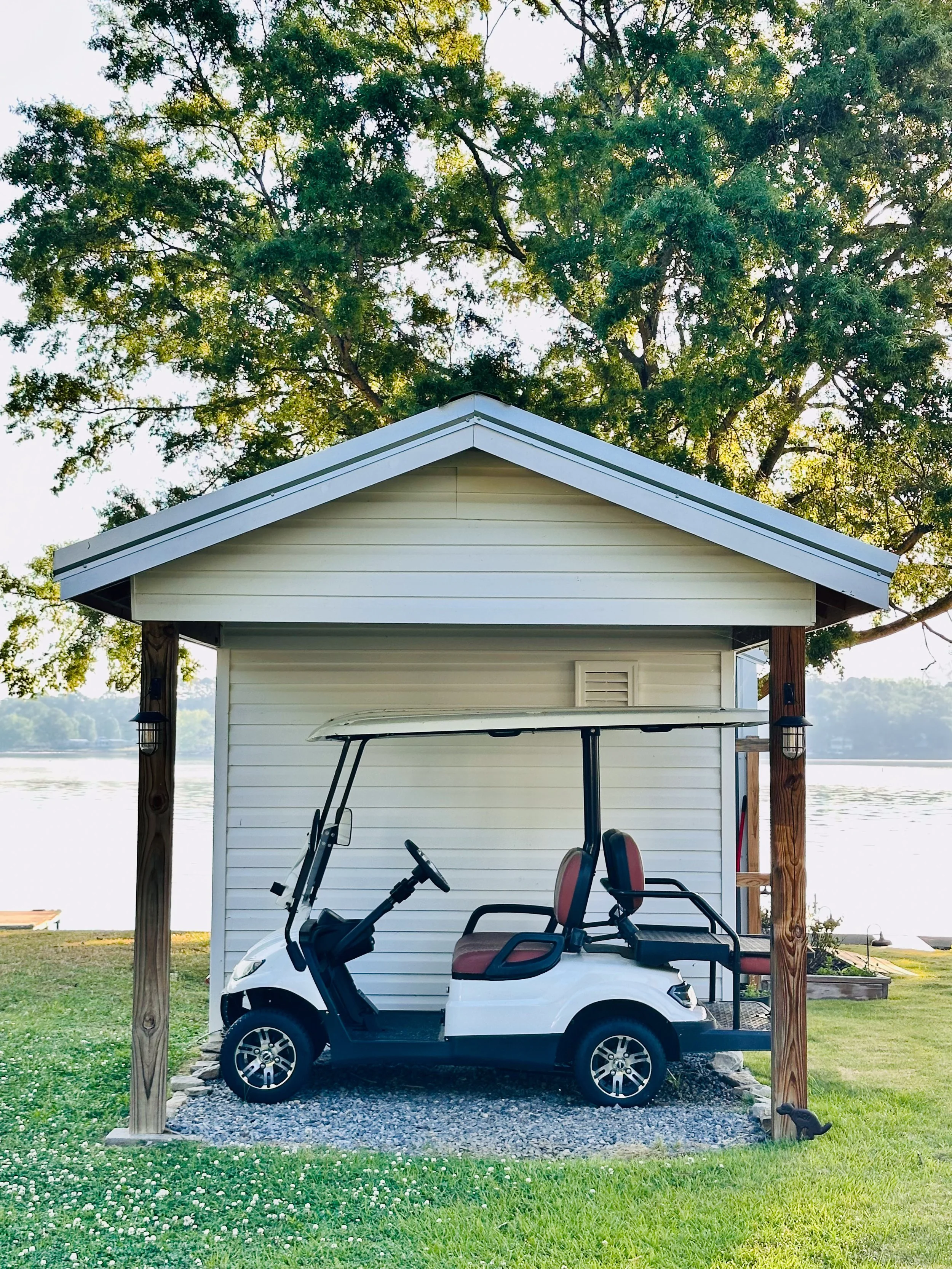 A white golf cart parked under a small wooden structure with a white roof, beside a white shed near a body of water, with trees and grassy area in the background.