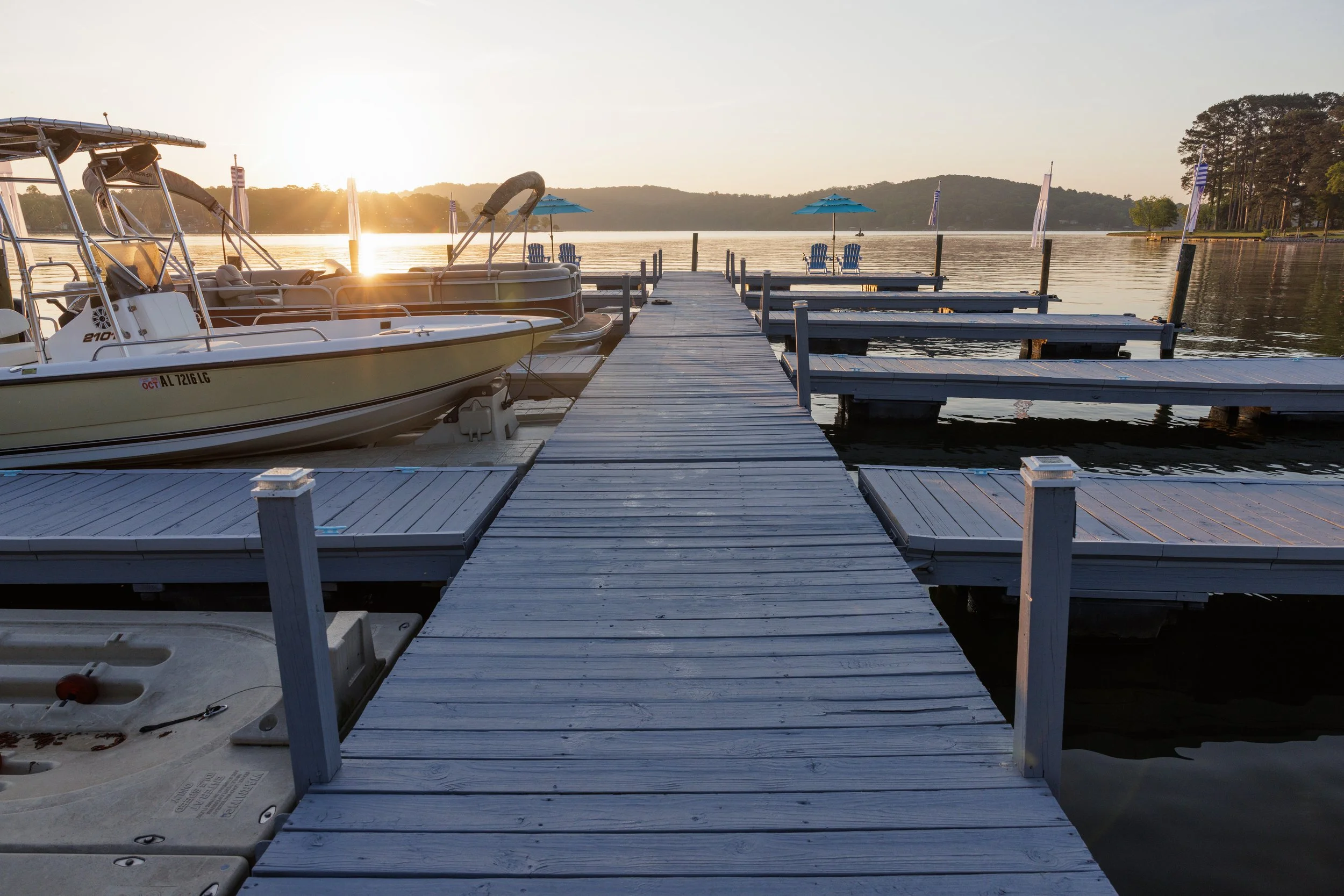 A dock extending into a calm lake with boats moored on the side and Adirondack chairs on the shore, during sunset with a hilly landscape in the background.