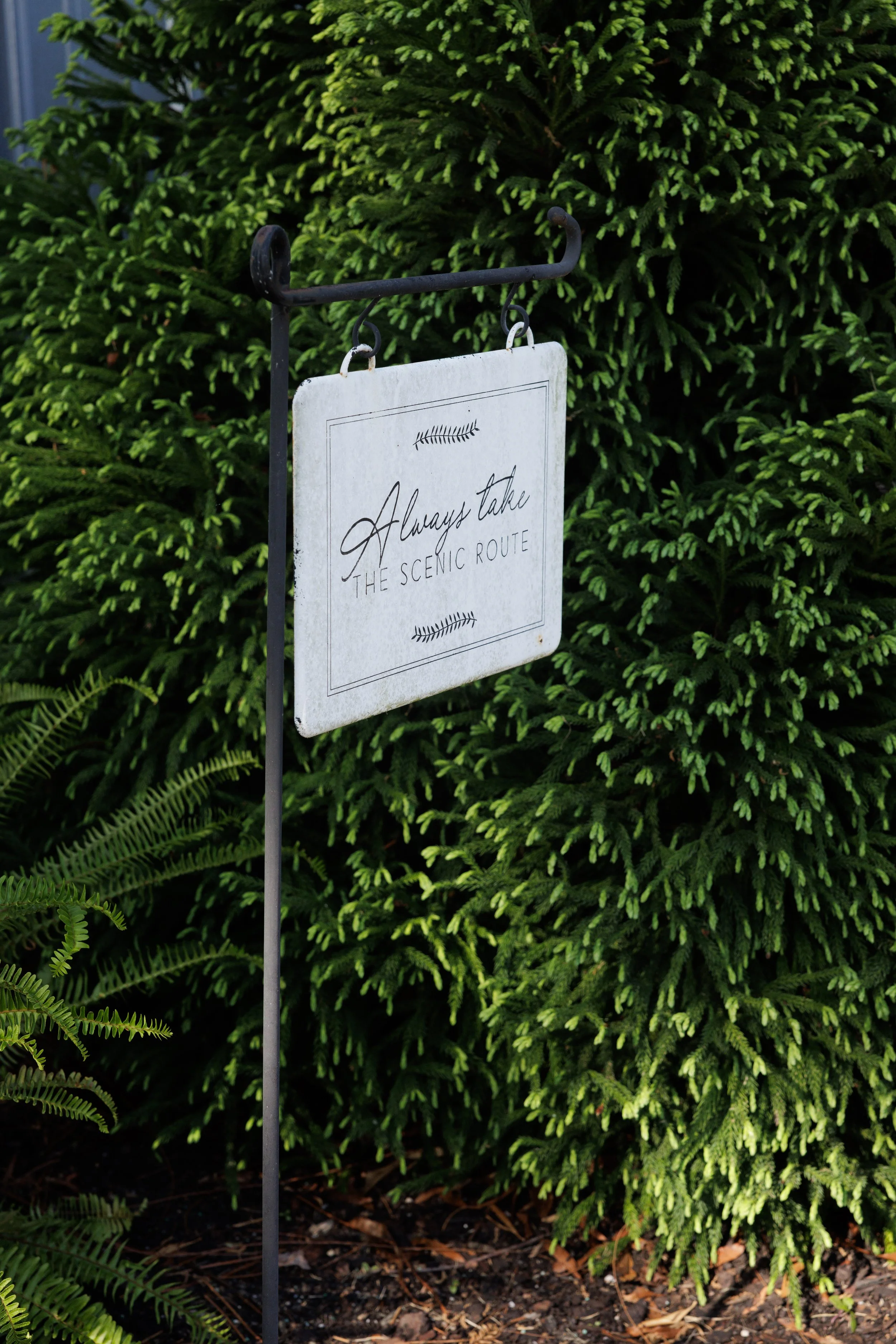 A decorative garden sign hanging from a black metal hook in front of lush green bushes, containing a quote that reads "Always take the scenic route" with small fern illustrations.