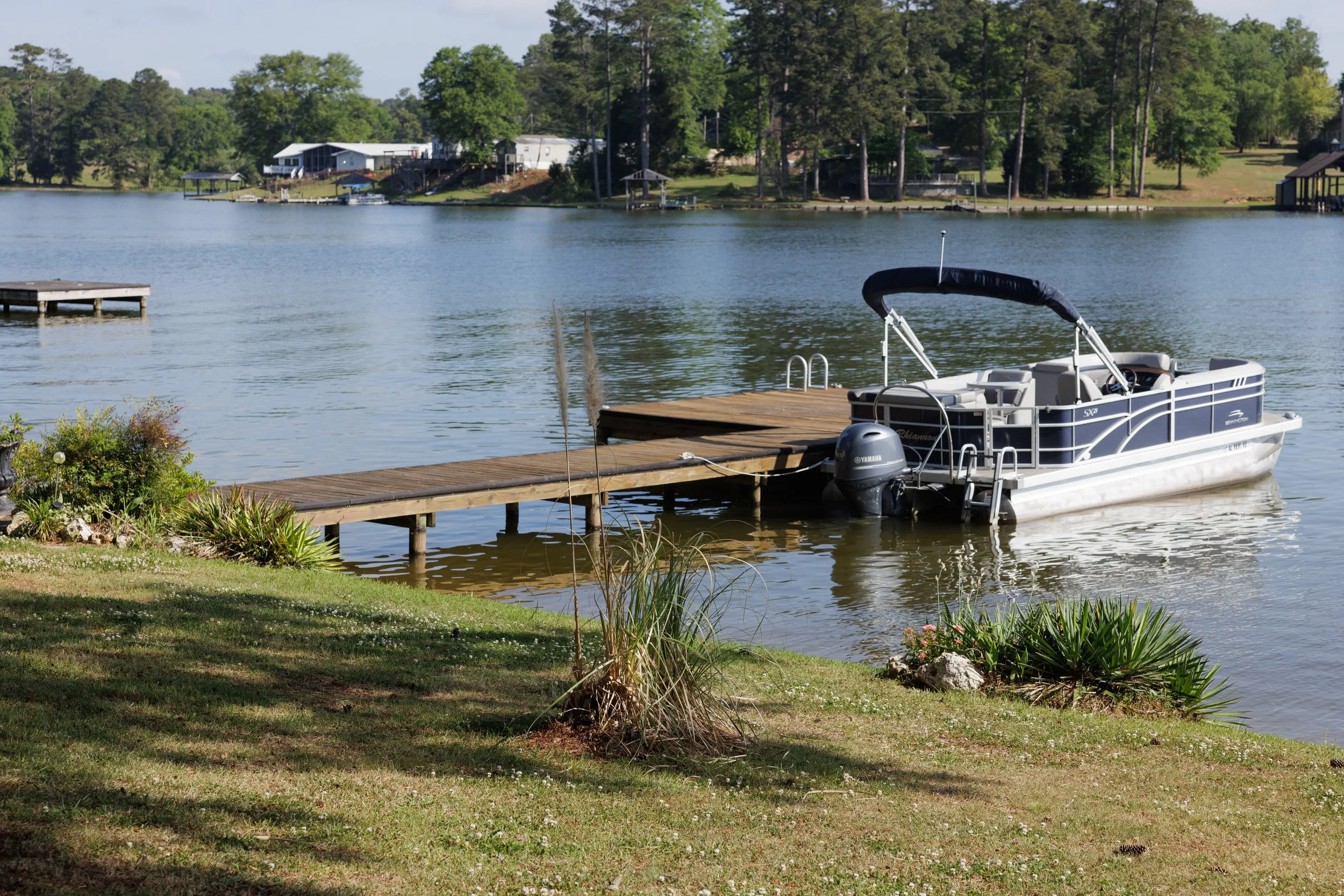 A boat docked at a wooden pier on a calm lake with houses and trees in the background.
