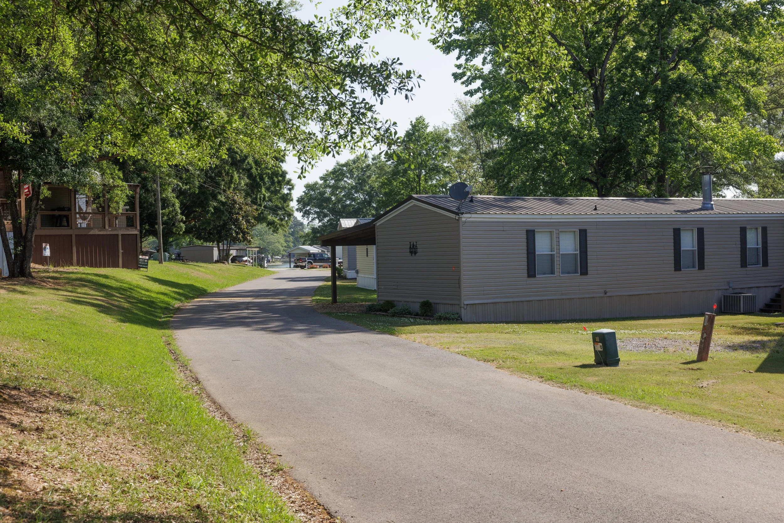 A residential area with a paved walkway, green grass, trees, mobile homes, and parked cars in the background on a sunny day.