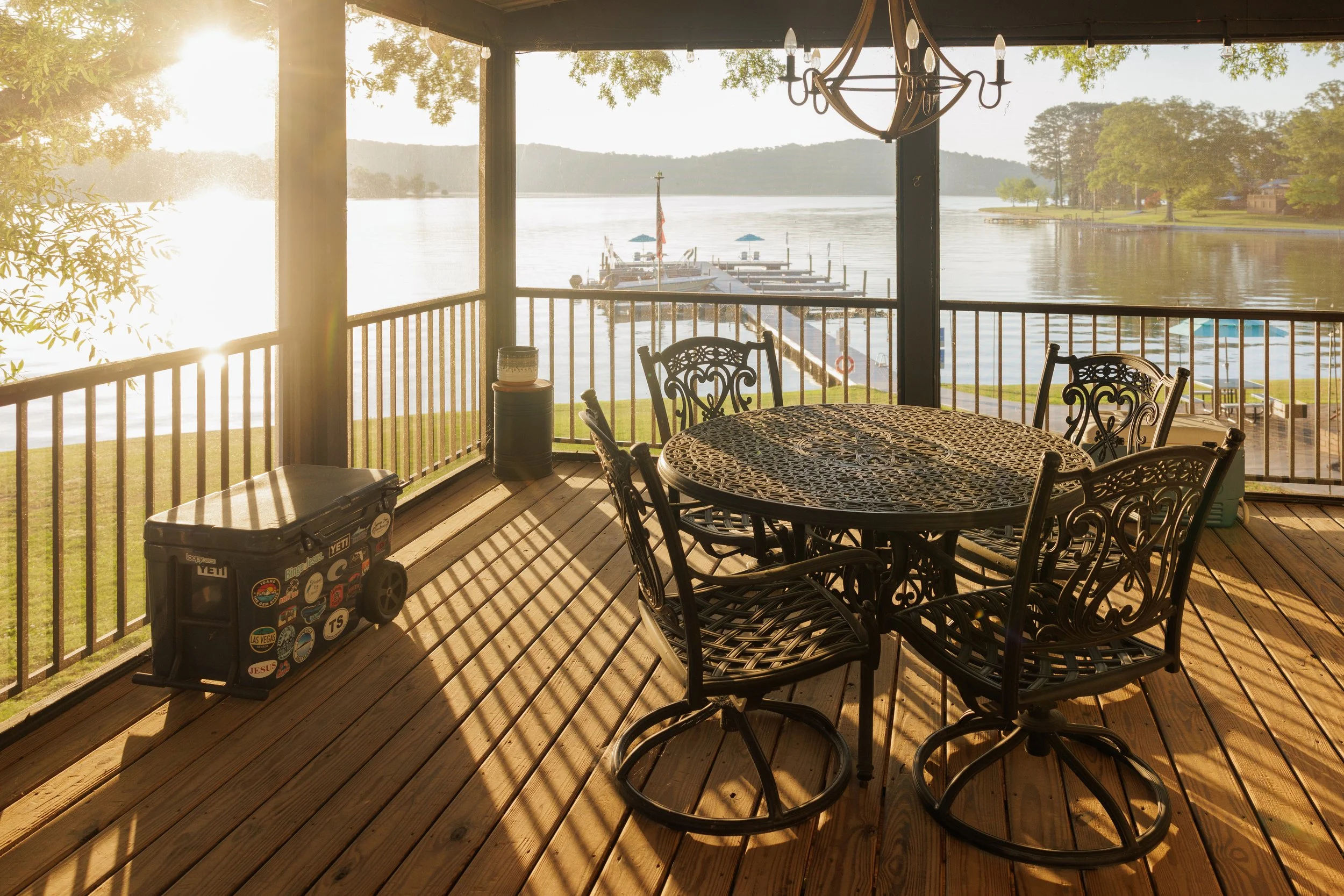 A lakeside porch with a round metal table and chairs, overlooking a docked boat on a calm lake, with trees and hills in the background, and the sun setting or rising, casting a warm glow.